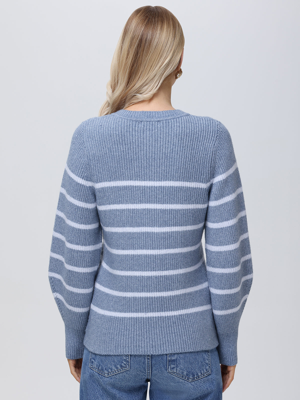 A person with long blonde hair, photographed from behind, wears a blue and white striped sweater by 525 America. The sweater is designed with balloon sleeves and solid cuffs, paired elegantly with blue jeans for a cozy yet sophisticated look.
