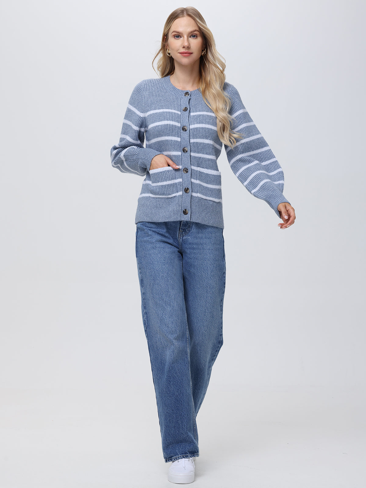 A person is relaxedly walking forward against a plain background, their hand in the pocket of their blue and white striped cardigan with large buttons and balloon sleeves by 525 America, paired with blue jeans.