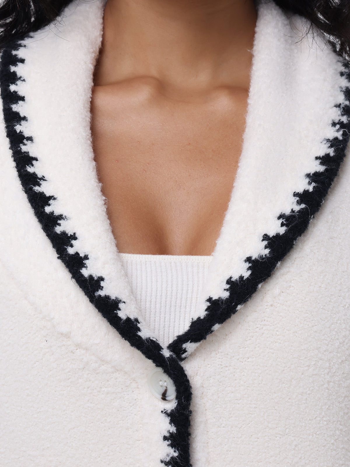 Close-up of the neckline of the 525 America Tierney Whipstitch Cardigan, highlighting white textured fabric with black zigzag trim. Its worn over a white ribbed top and features whipstitch detailing with a visible button.