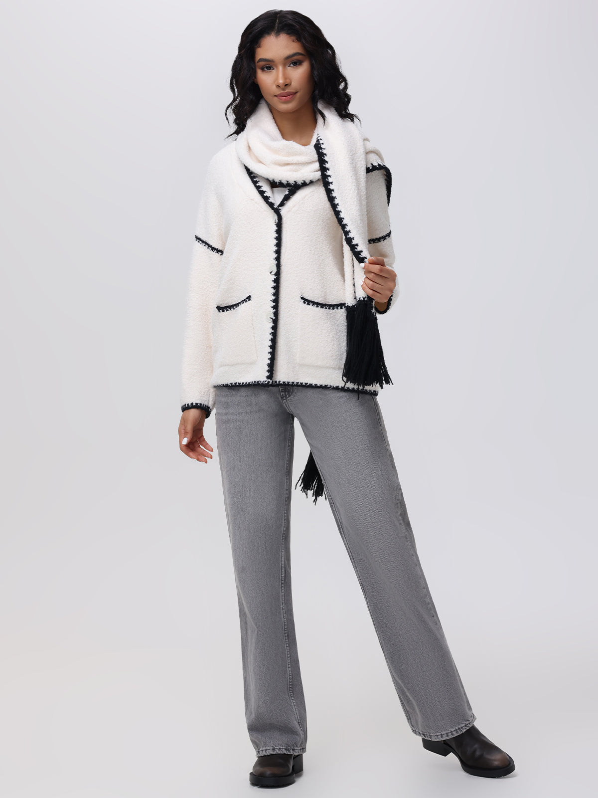 A person with long, dark hair stands against a neutral background wearing a white 525 America sweater with whipstitch details, paired with gray jeans and black shoes. One hand rests by their side while the other holds the removable scarf elegantly draped around their neck.