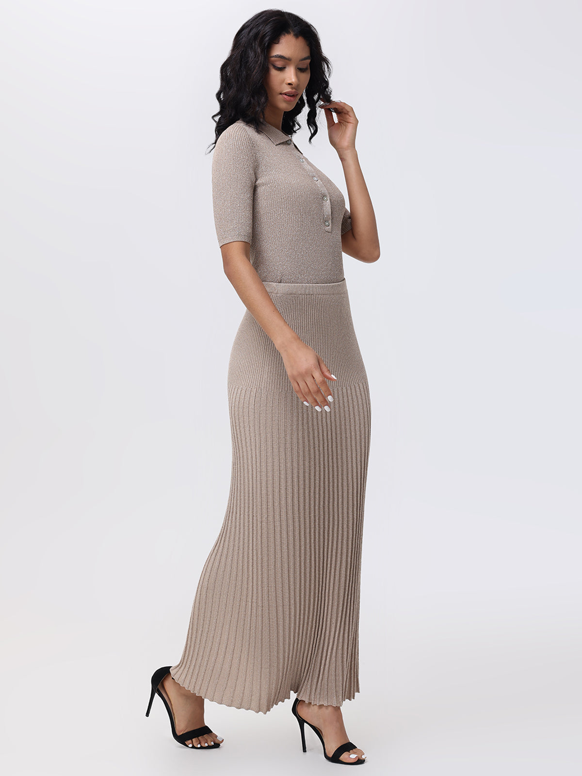 A woman with long dark hair wears a fitted, ribbed taupe dress with elegant pleats and a buttoned top from 525 America. She stands sideways, holding the collar, wearing black high-heeled sandals against a plain light gray background.