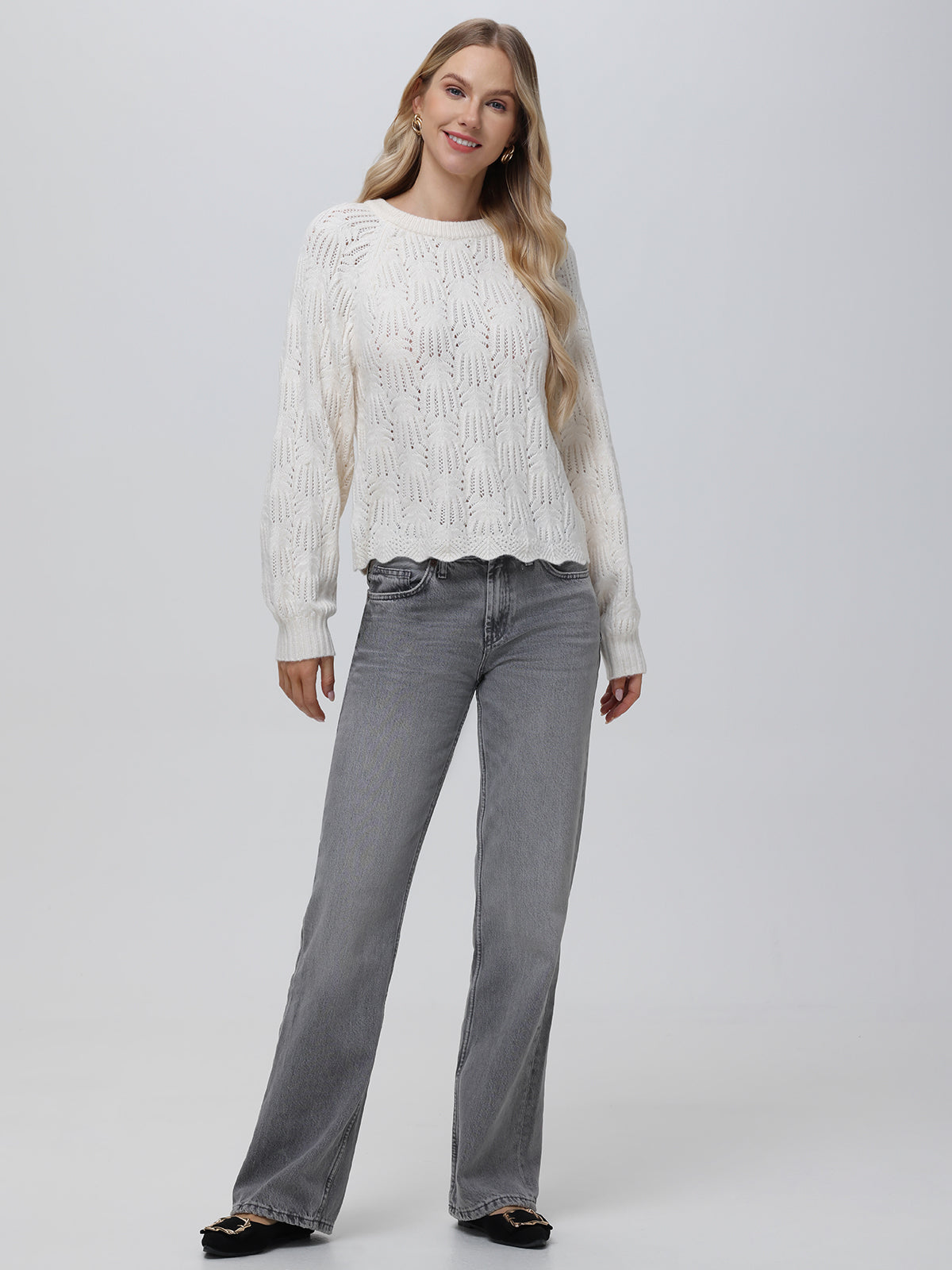 A person with long, wavy hair smiles against a plain background in a white 525 America pullover featuring pointelle detailing and a crew neckline, paired with gray jeans and black shoes. Their hands rest at their sides, giving off a relaxed vibe.