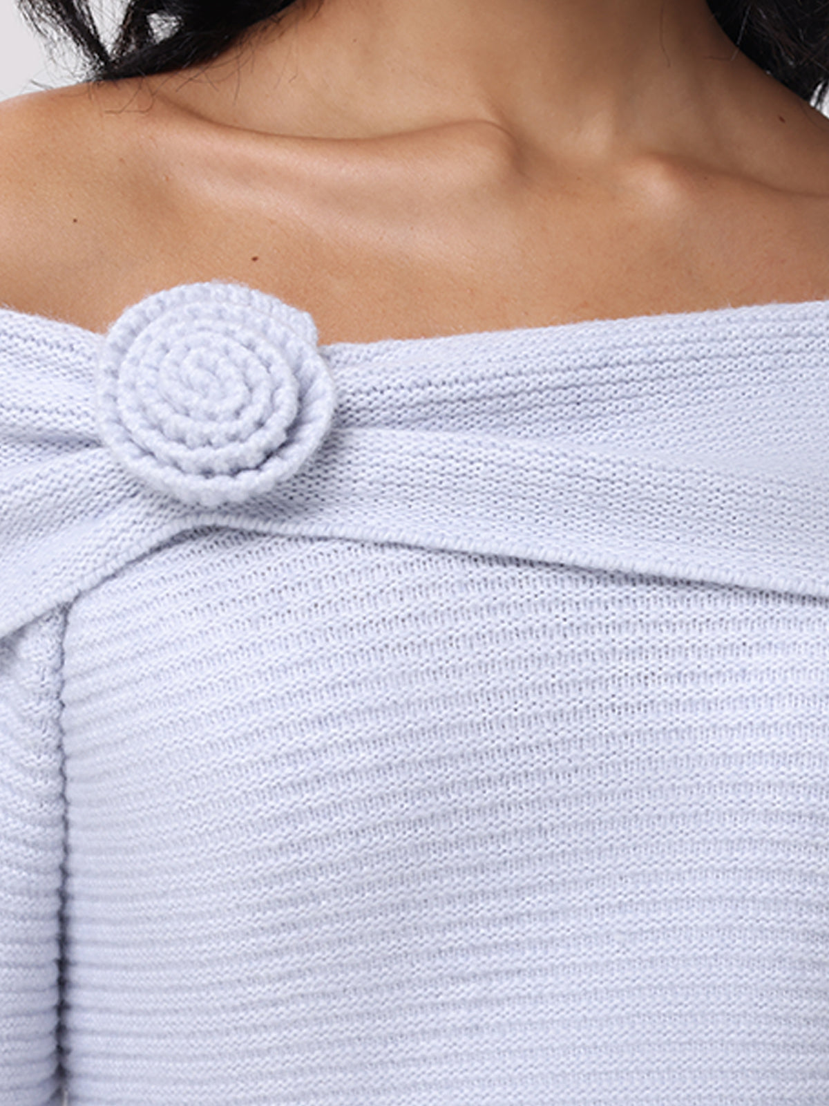 A close-up showcases someone wearing 525 Americas Elizabeth Sweater, featuring a light gray off-the-shoulder design with ribbed texture and a rosette accent at the neckline, highlighting the shoulders and collarbone.