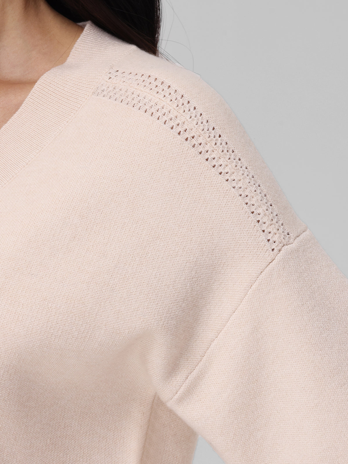 Close-up of a persons shoulder in a 525 America light beige knitted jumper. The sweater showcases timeless elegance with its subtle filigree openwork along the shoulder seam against a neutral gray background.