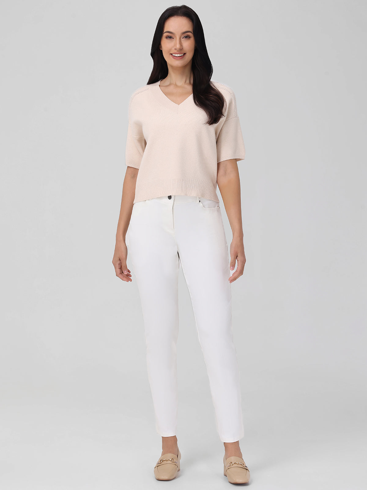 A woman, smiling directly at the camera, stands against a gray backdrop in a 525 America light beige V-neck short-sleeve sweater paired with white pants and beige loafers. Her long dark hair adds to the timeless elegance of her look.