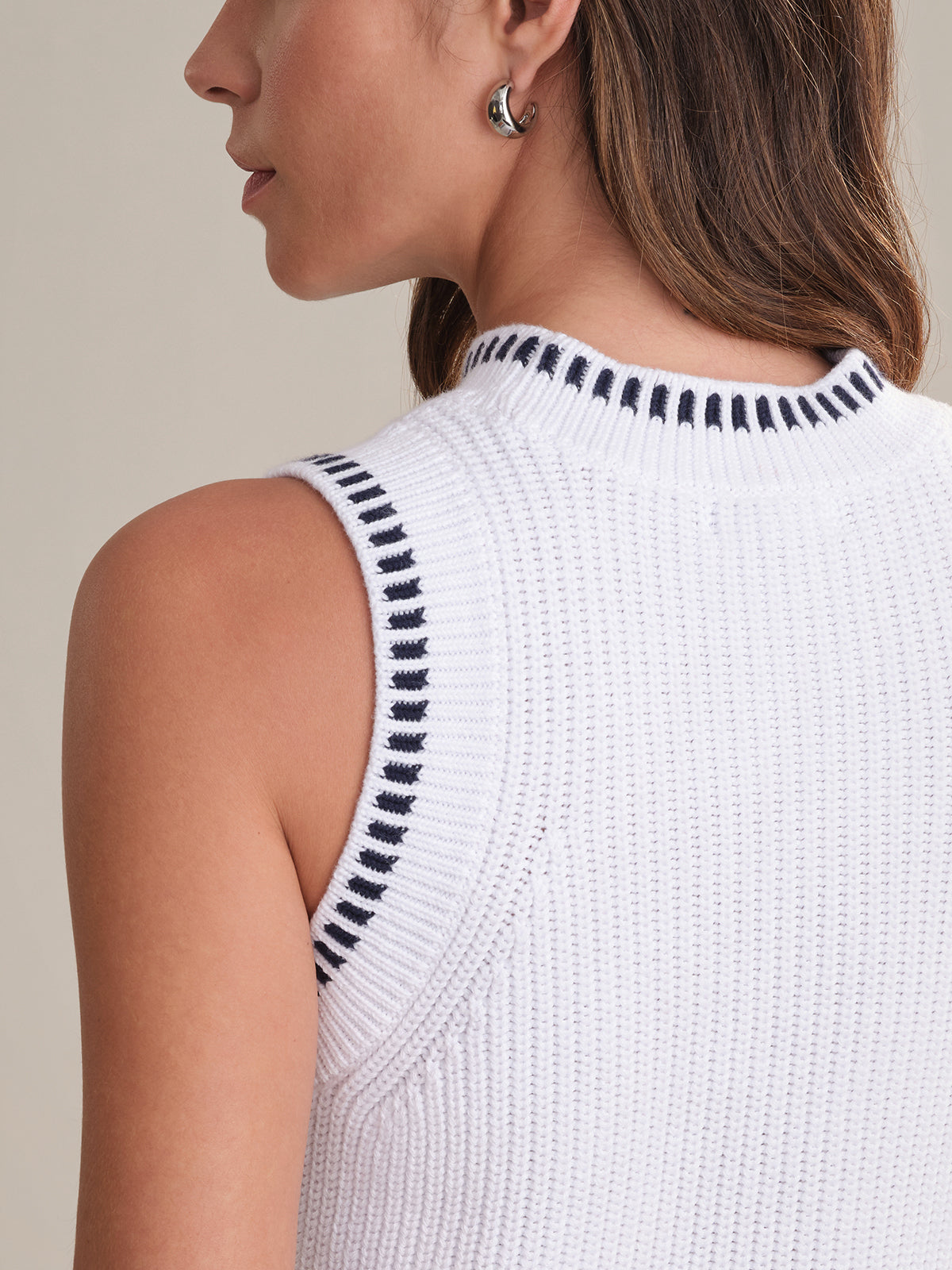 A woman with long brown hair and a silver hoop earring models the 525 America sleeveless white cotton sweater-tank with black trim at the armholes and collar, shown from the back and side.