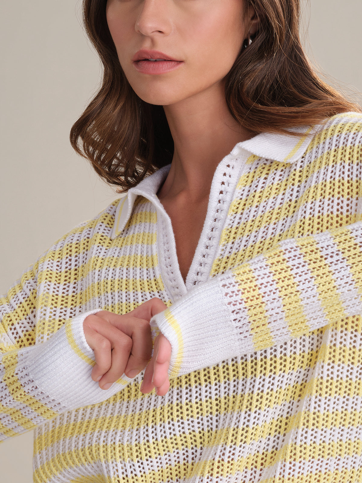 A woman models the 525 America Kimberly Stripe Polo Sweater, made from 100% cotton with a collared v-neck. Shown shoulders up, her right arm is bent and her hand emerges slightly from the sleeve’s thumb hole.