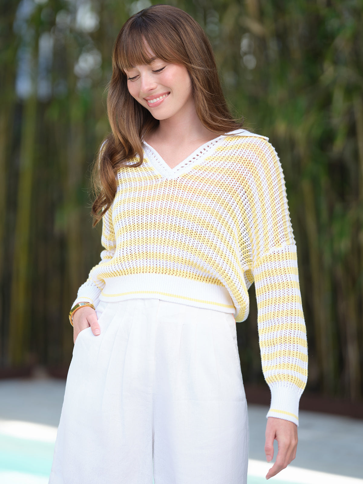 A woman with long brown hair smiles while looking down. She is wearing a 525 America 100% cotton Kimberly Stripe polo sweater and white pants, standing outdoors with green foliage in the background.