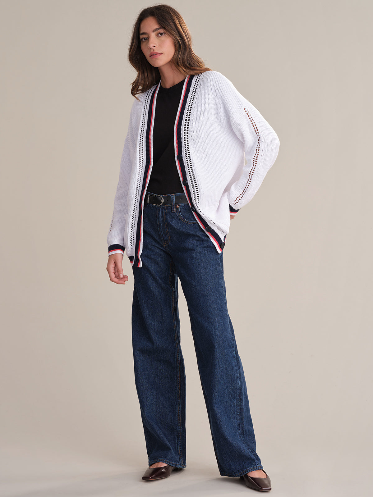 A person stands against a plain background wearing a 525 America relaxed fit cotton cardigan in varsity style with red and black trim, paired with a black shirt, blue wide-leg jeans, and brown shoes.