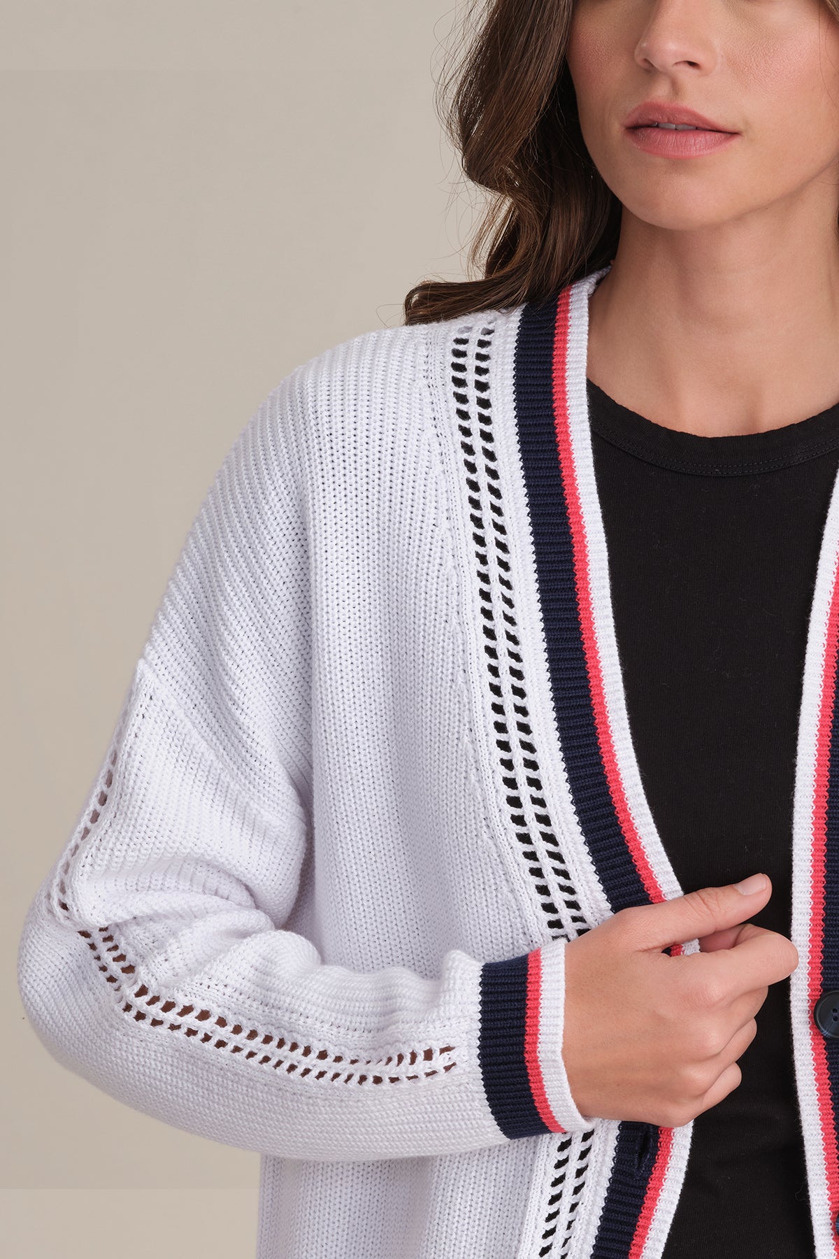 A woman models a 525 America relaxed fit white cotton cardigan with varsity-style navy and red trim over a black top, holding the cardigan. Her lower face, neck, and upper torso appear against a plain beige background.