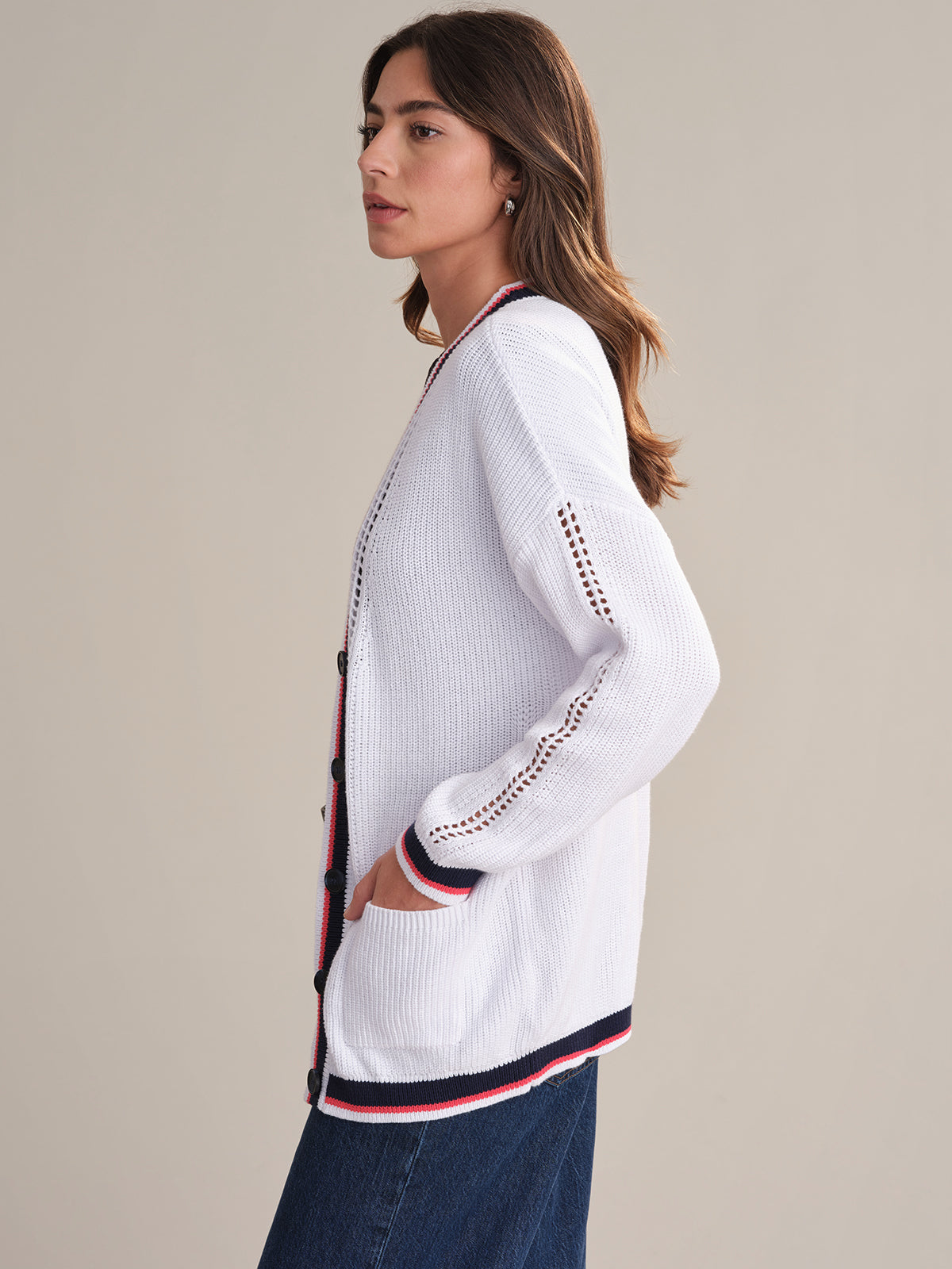 A woman with long brown hair stands in profile against a plain light background, wearing a 525 America relaxed fit varsity cotton cardigan with red and navy trim, eyelet sleeve details, and blue jeans.