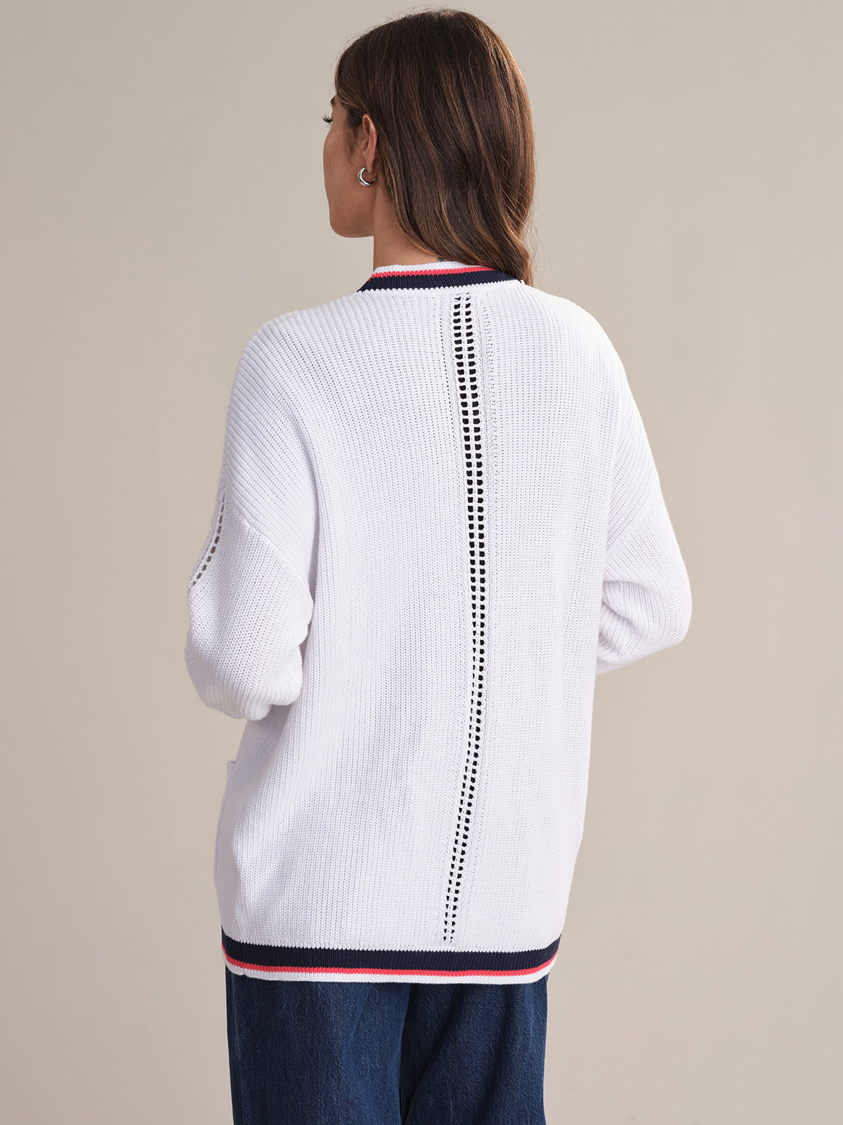 A person with medium-length brown hair is seen from behind wearing a 525 America relaxed fit cotton cardigan featuring decorative vertical openwork on the back and red, white, and black varsity-style trim on the collar and hem.