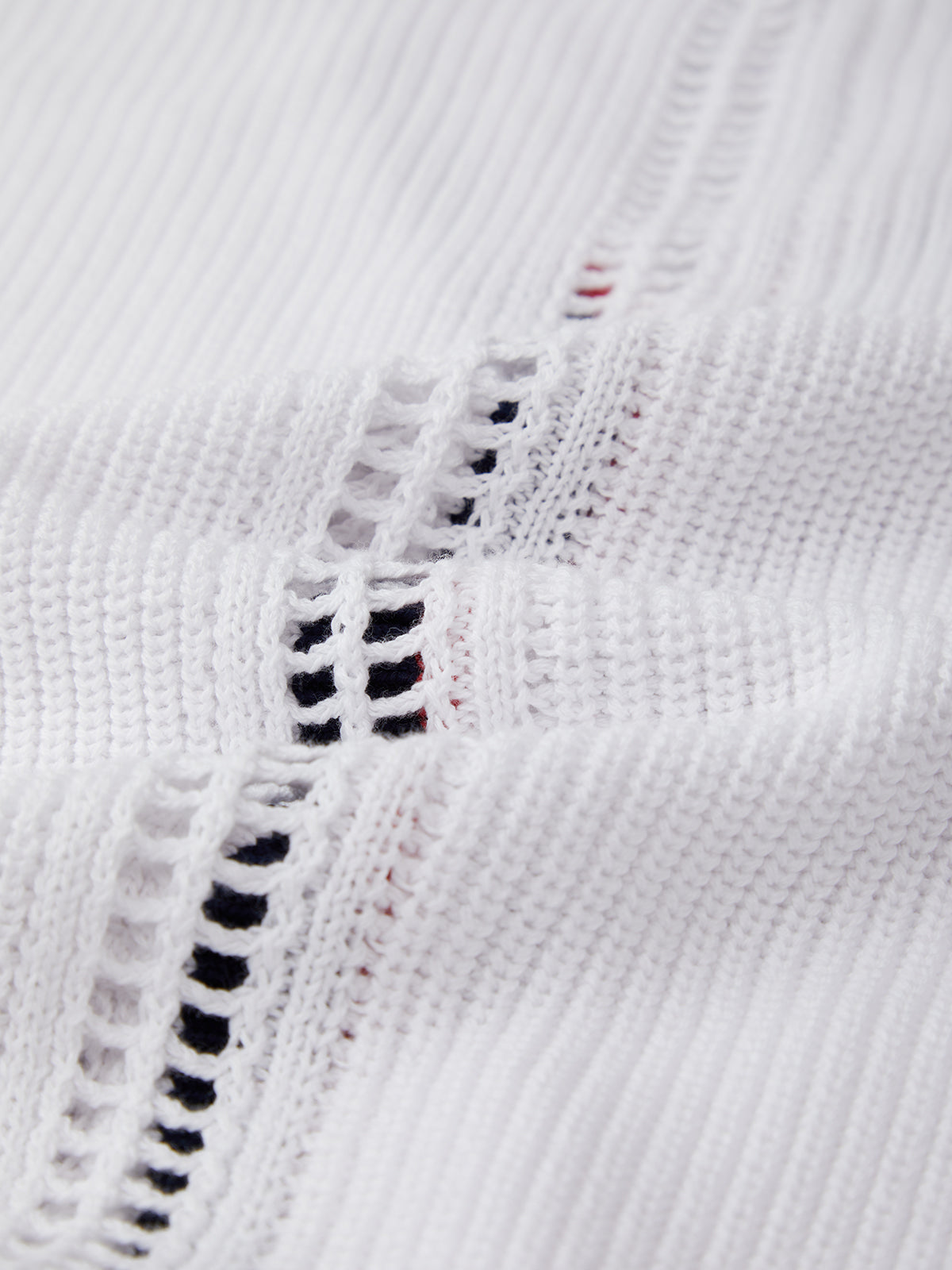 Close-up of a 525 America white knitted cotton cardigan with horizontal openwork rows, revealing black and red threads underneath for a varsity-inspired look. The knit’s texture and pattern are clearly displayed.