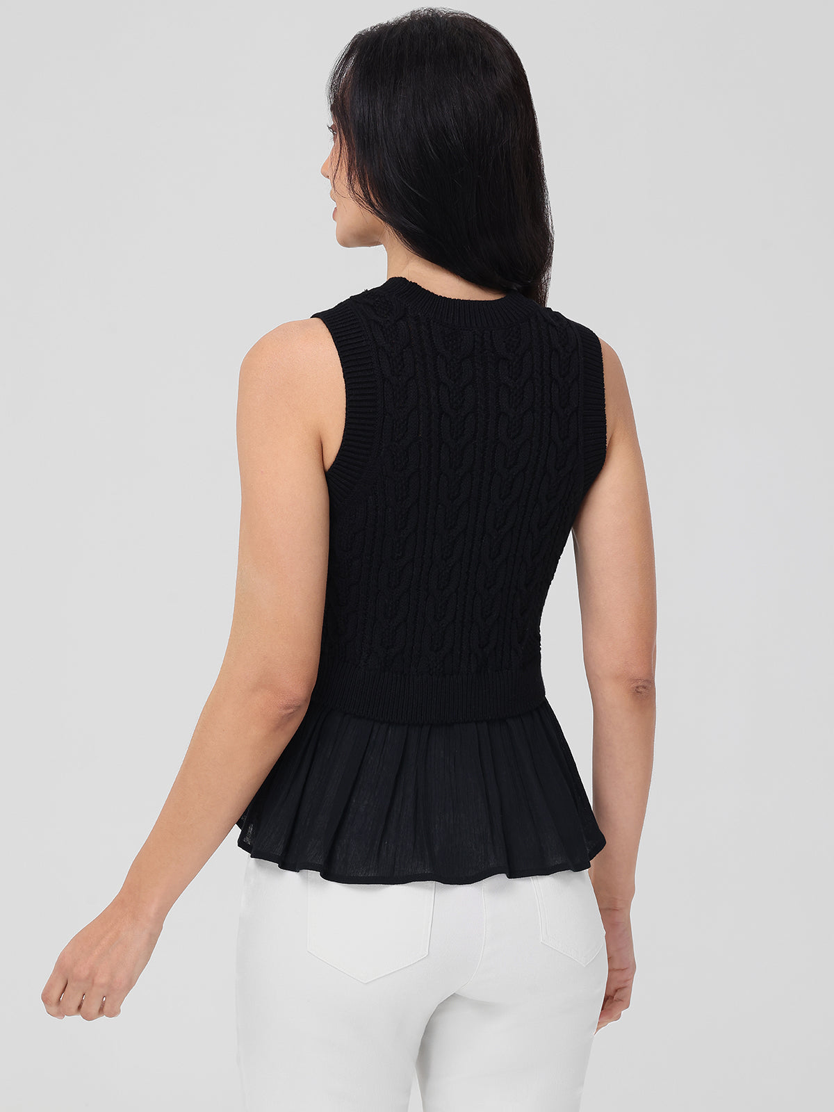 Rear view of a person with long dark hair wearing a 525 America black sleeveless cable-knit sweater featuring a peplum bottom, made from 100% cotton and ideal for layering. It is paired with white pants against a plain light gray background.