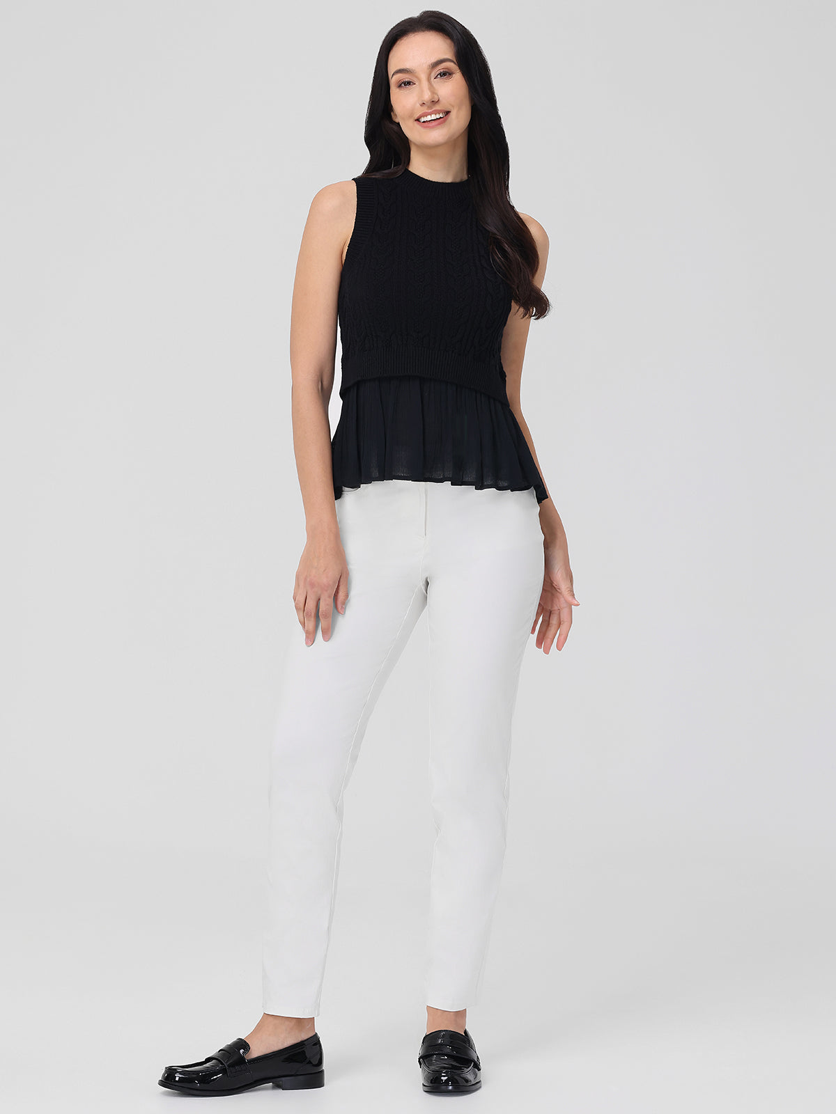 A person with long dark hair stands against a plain background, wearing a sleeveless black top by 525 America and white 100% cotton pants, paired with black loafers. They smile warmly, one hand on their thigh and the other slightly raised.