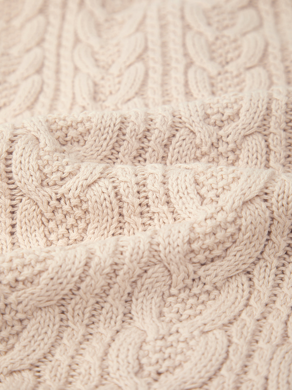 Close-up of a cream-colored, cable-knit fabric by 525 America. Made from 100% cotton with a prominent braided pattern, its soft texture offers cozy comfort and versatility for layering in any season.
