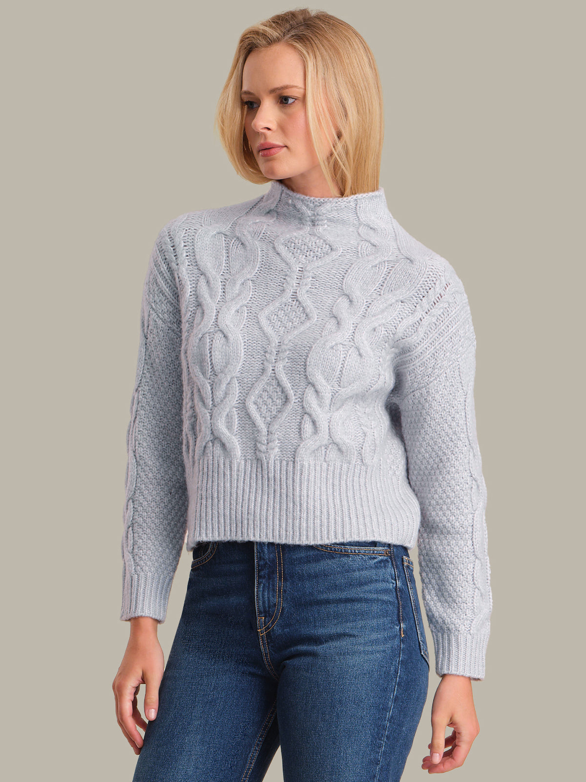 Woman wearing a light blue cable knit sweater and blue jeans against a plain background