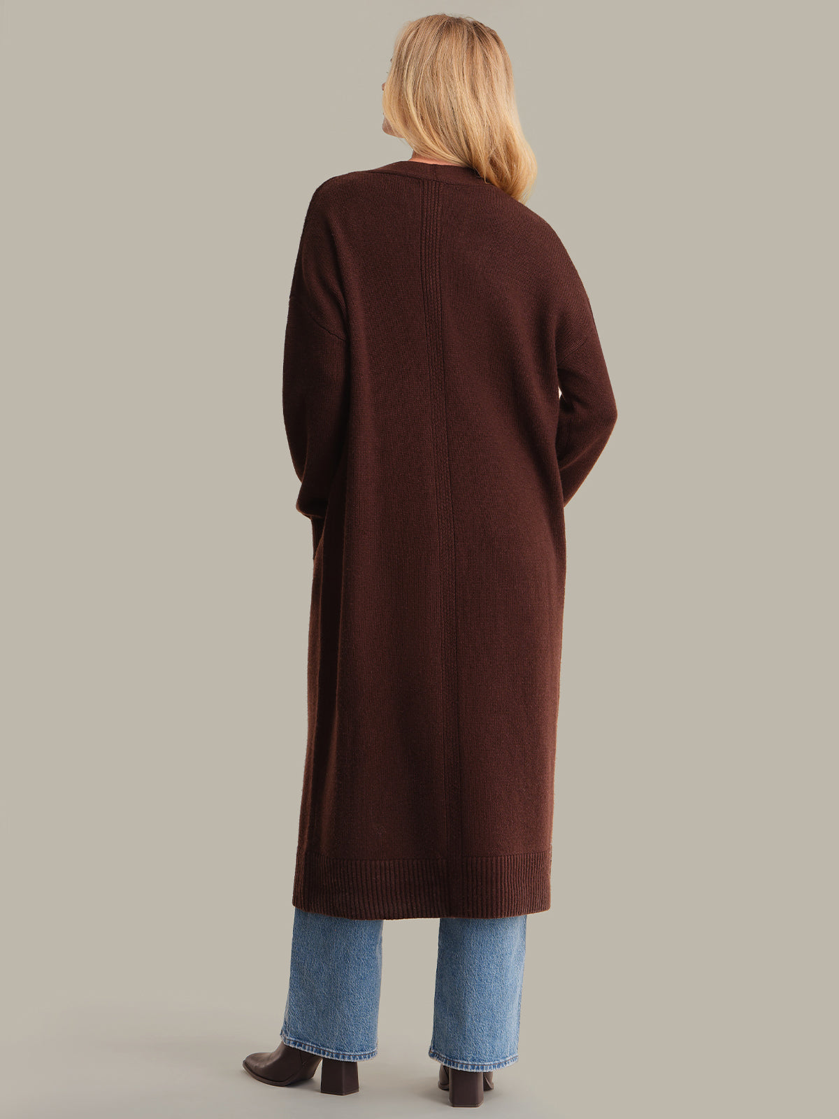 Person wearing a long brown cardigan and blue jeans on a plain background