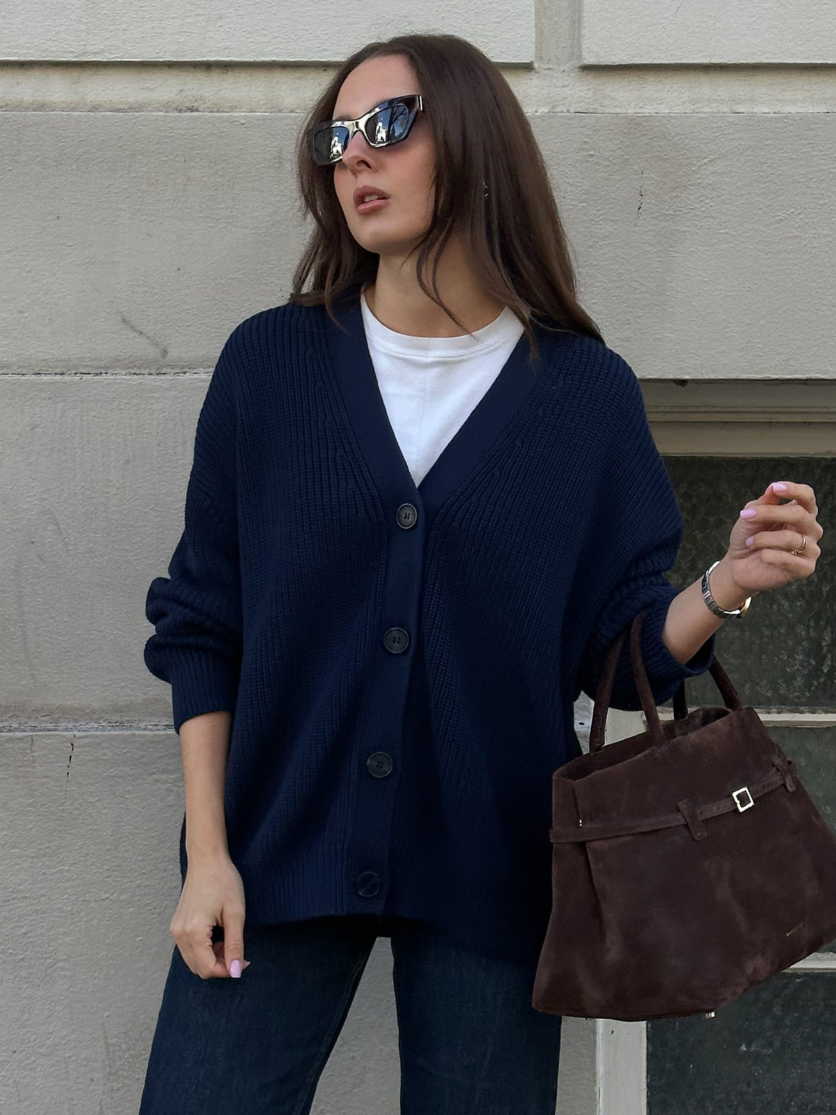 A woman with long brown hair and sunglasses wears a navy blue Emilia: Cotton Cocoon Cardigan over an oversized 100% cotton white top. She pairs it with dark jeans and carries a brown suede handbag, striking a pose against a light-colored wall.