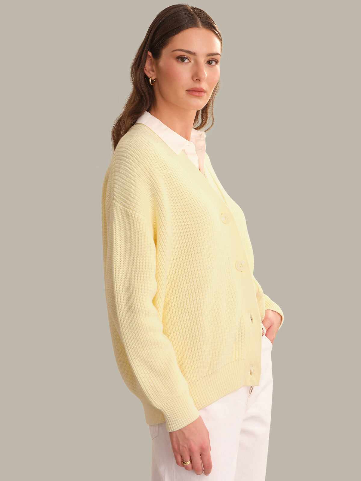 A woman wearing a pale yellow ribbed knit cardigan over a white collared shirt and white pants, photographed against a plain light gray studio background.