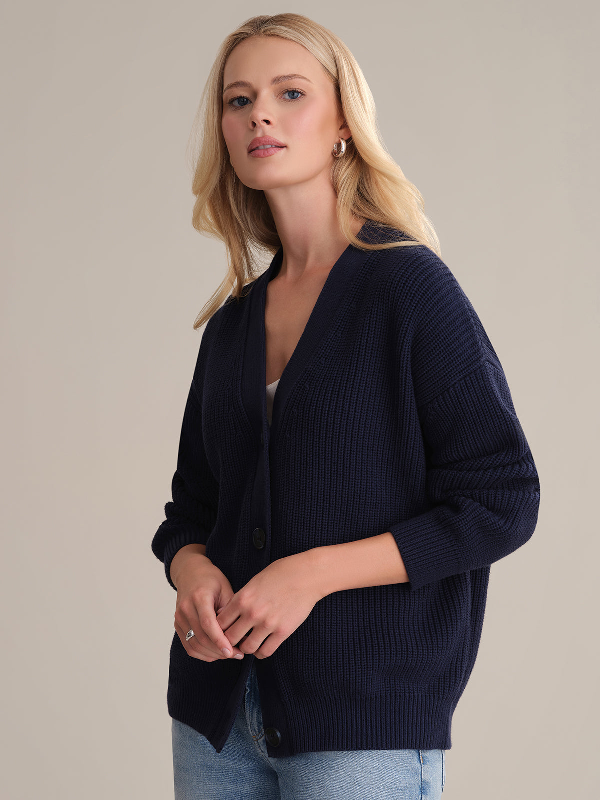 A person with long, blonde hair wears the Emilia: Cotton Cocoon Cardigan in an oversized dark blue style, made from 100% cotton, over a white top. They stand against a neutral background, calm expression, hands resting on the cardigans sides.