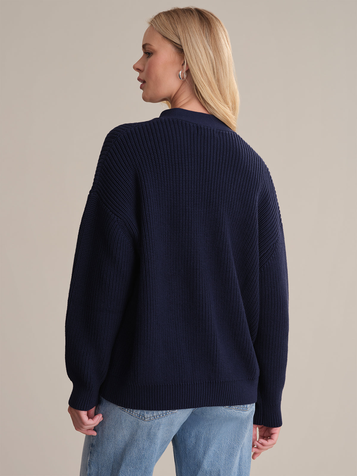 A person with long blonde hair is shown from the back, wearing a 525 America navy blue oversized knit sweater and light blue jeans, standing against a plain beige background.