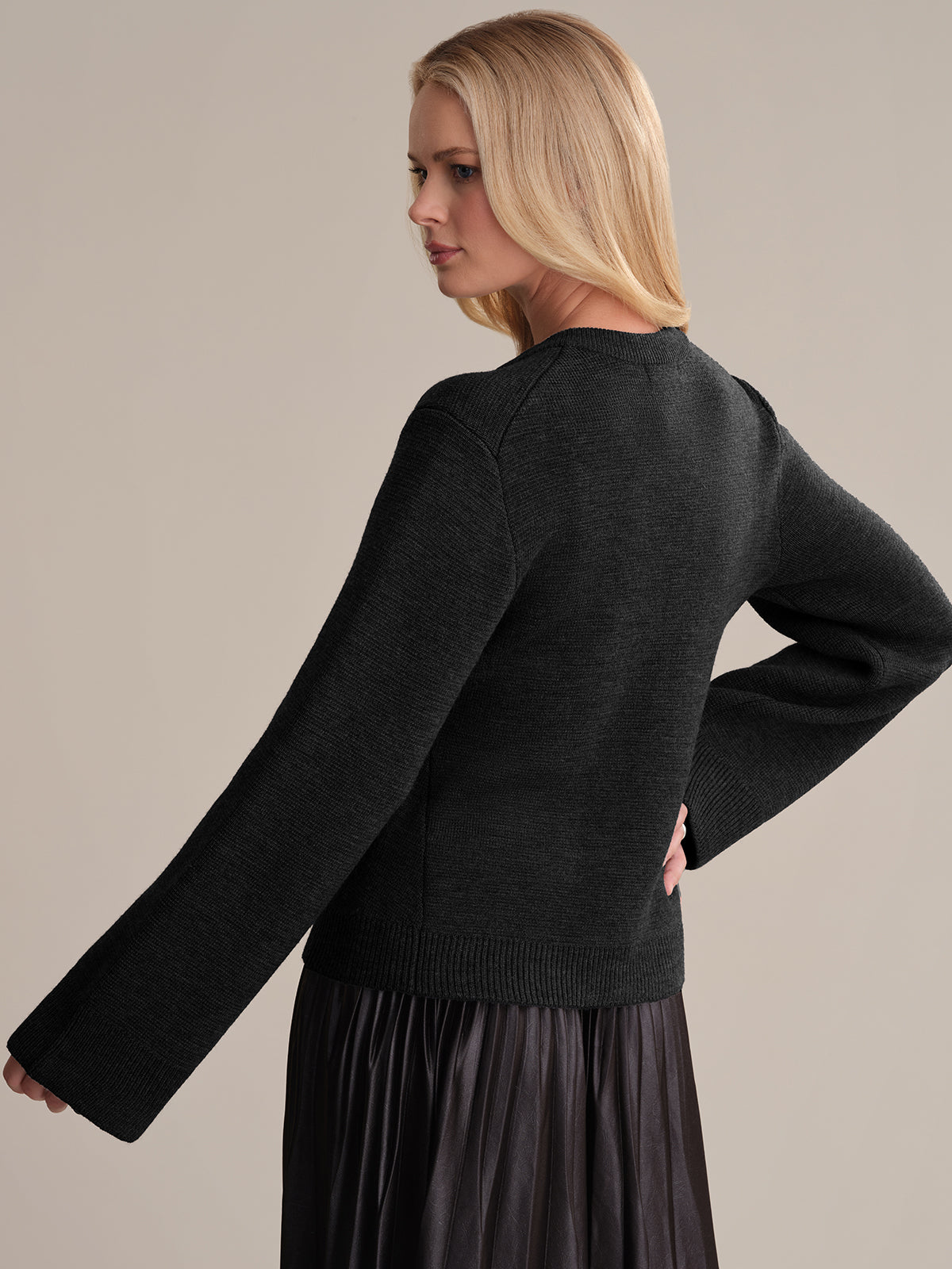 A person with long blonde hair radiates sophistication in a black knit sweater over a pleated skirt, confidently posing with one hand on their hip against a neutral backdrop, embodying elegance akin to the Catherine: Wool Cardigan.