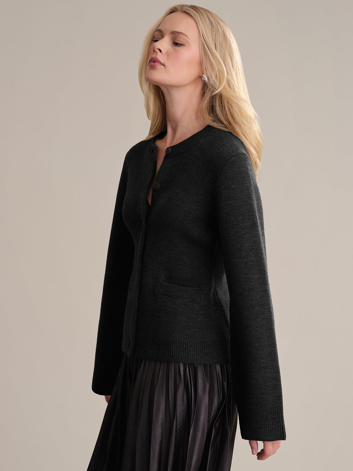 With eyes closed, a person with long blonde hair exudes sophistication in the Catherine: Wool Cardigan paired with a long pleated skirt. The plain beige background complements their elegant profile.