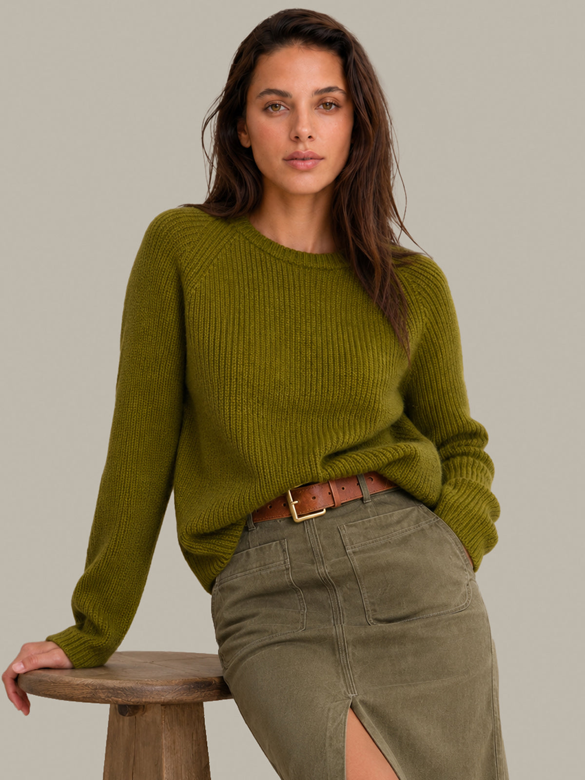 A woman wearing a ribbed olive green long-sleeve sweater and an olive green skirt, leaning on a wooden stool against a solid light gray background.