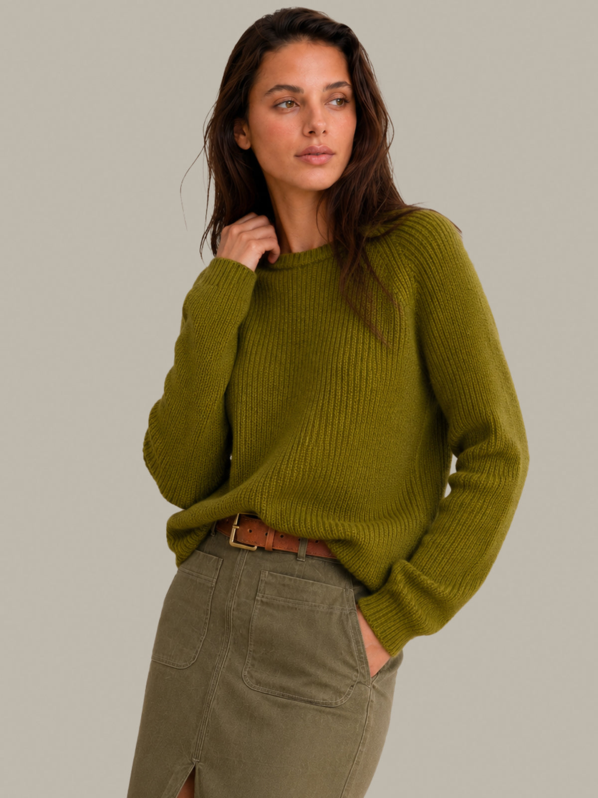 A woman wearing a ribbed knit crewneck sweater in olive green, paired with an olive green skirt and a brown belt, against a solid light gray studio background.