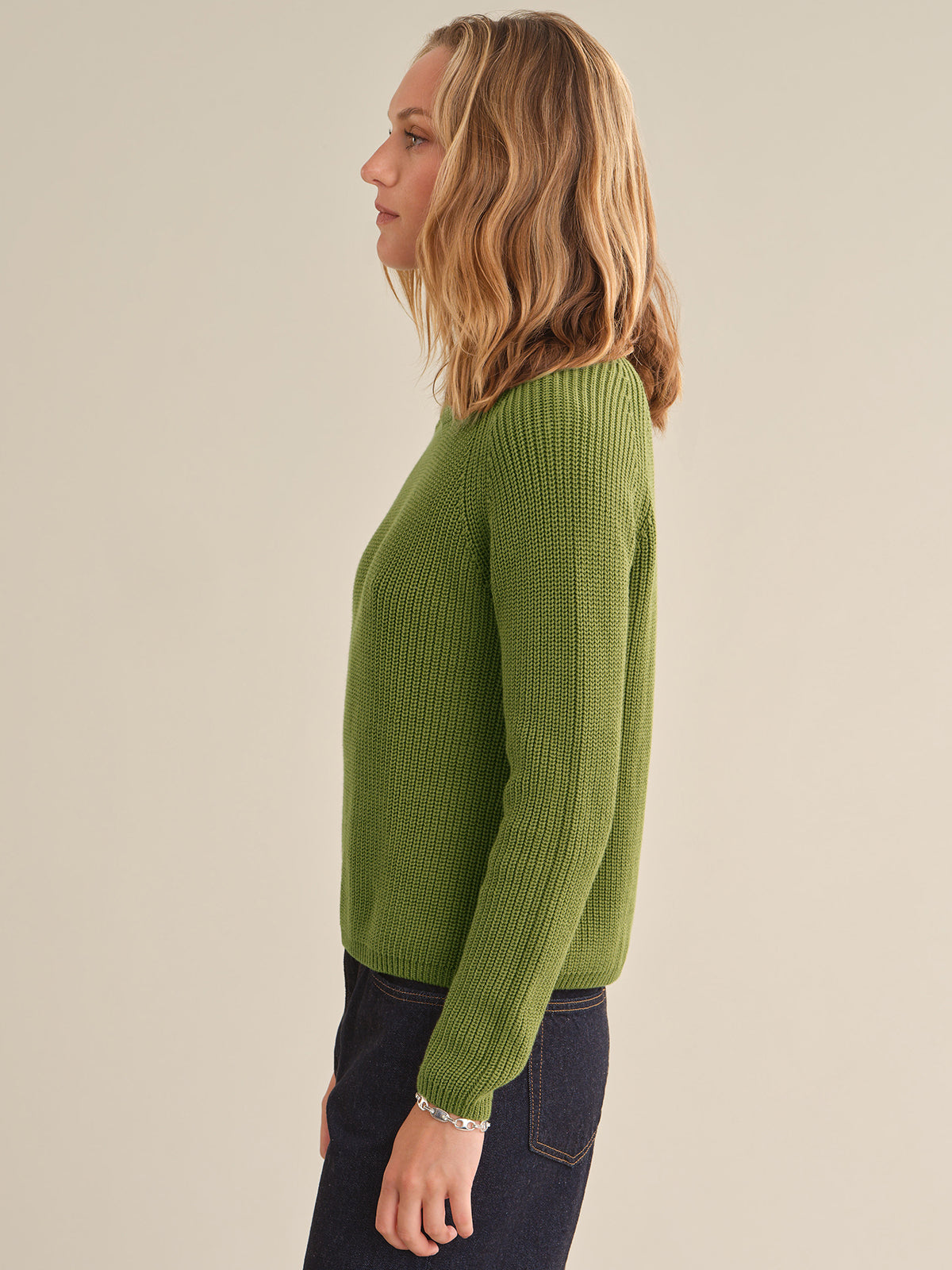 A woman shown in side profile wearing a ribbed knit long-sleeve sweater in olive green, paired with dark jeans, against a plain light beige studio background.
