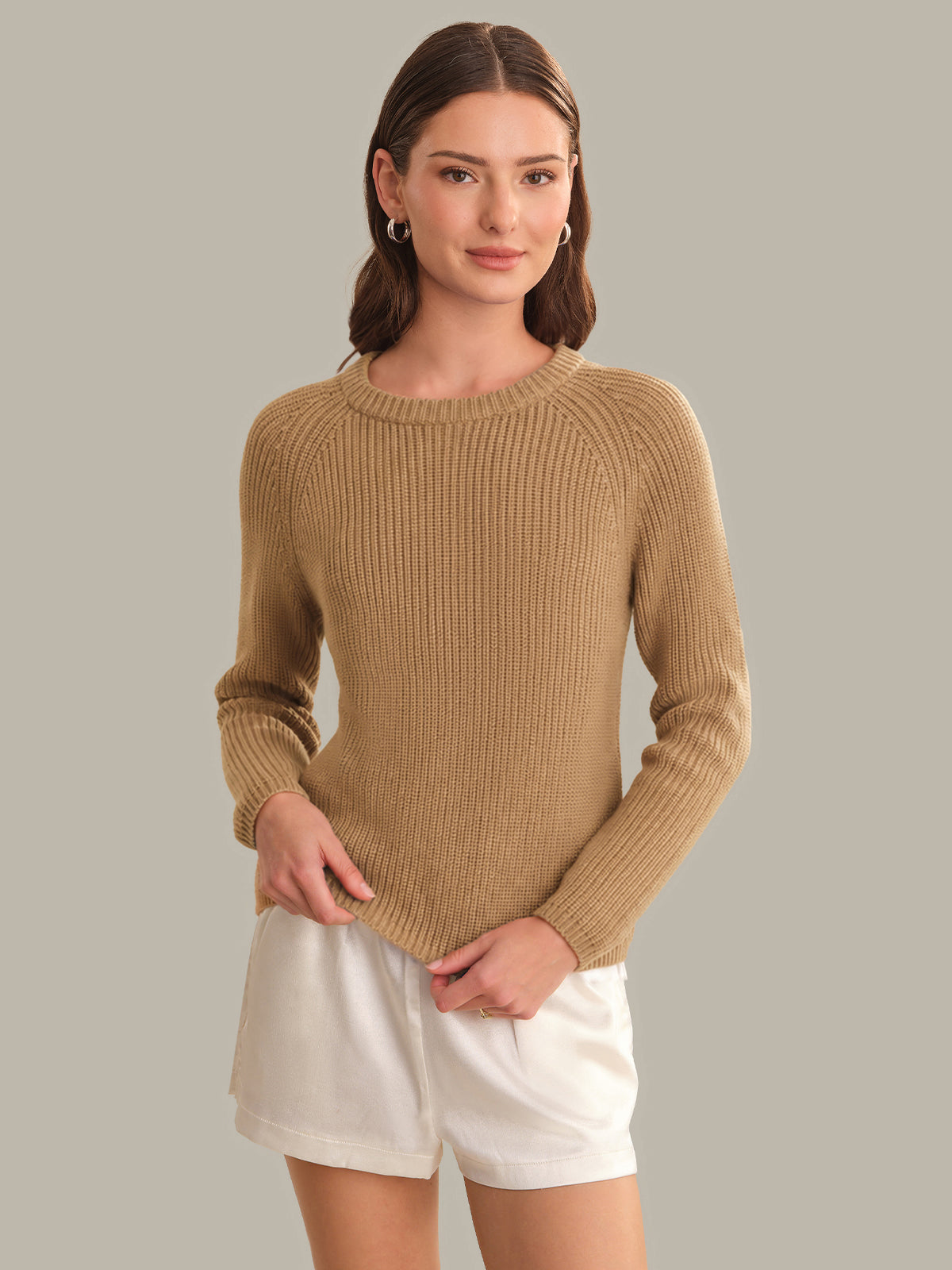 Woman wearing a beige sweater and white shorts against a gray background