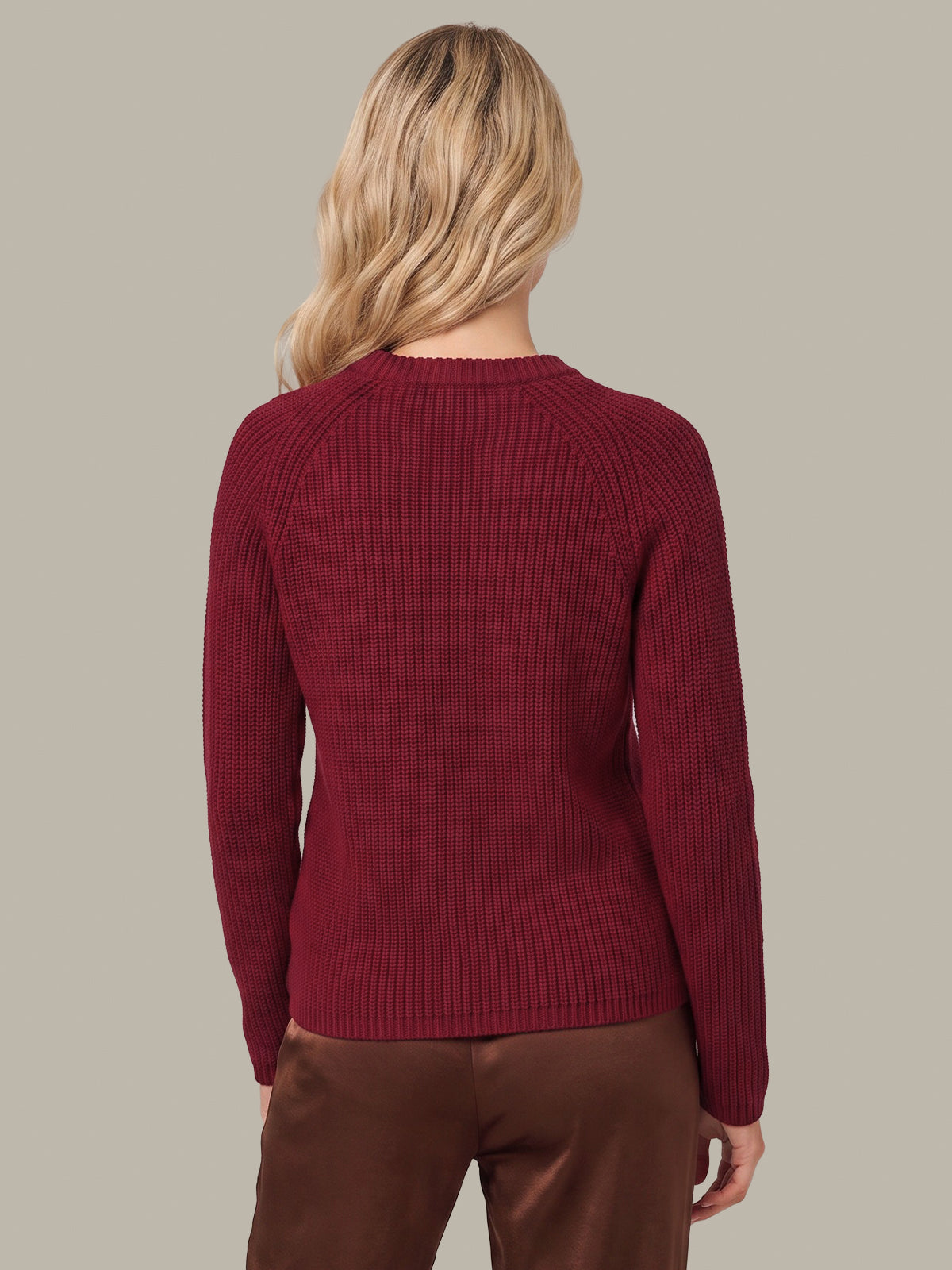 A woman viewed from the back wearing a ribbed knit long-sleeve sweater in deep burgundy and brown pants, set against a solid light gray background.