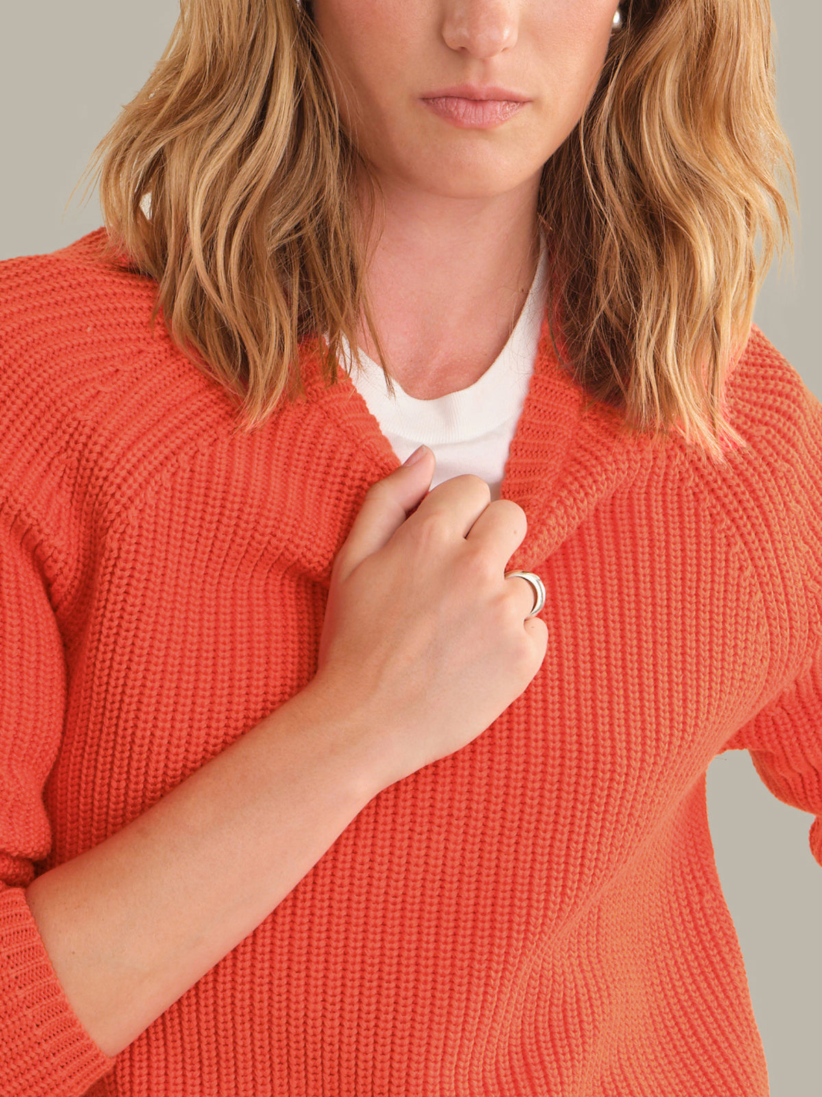 A close-up of a woman wearing a textured orange knit cardigan over a white t-shirt, with her hand resting on the front of the cardigan.