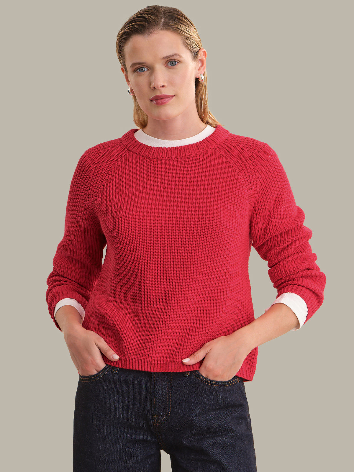 A woman wearing a red ribbed knit crewneck sweater with white contrast collar and cuffs, paired with dark blue jeans, against a solid light gray background.