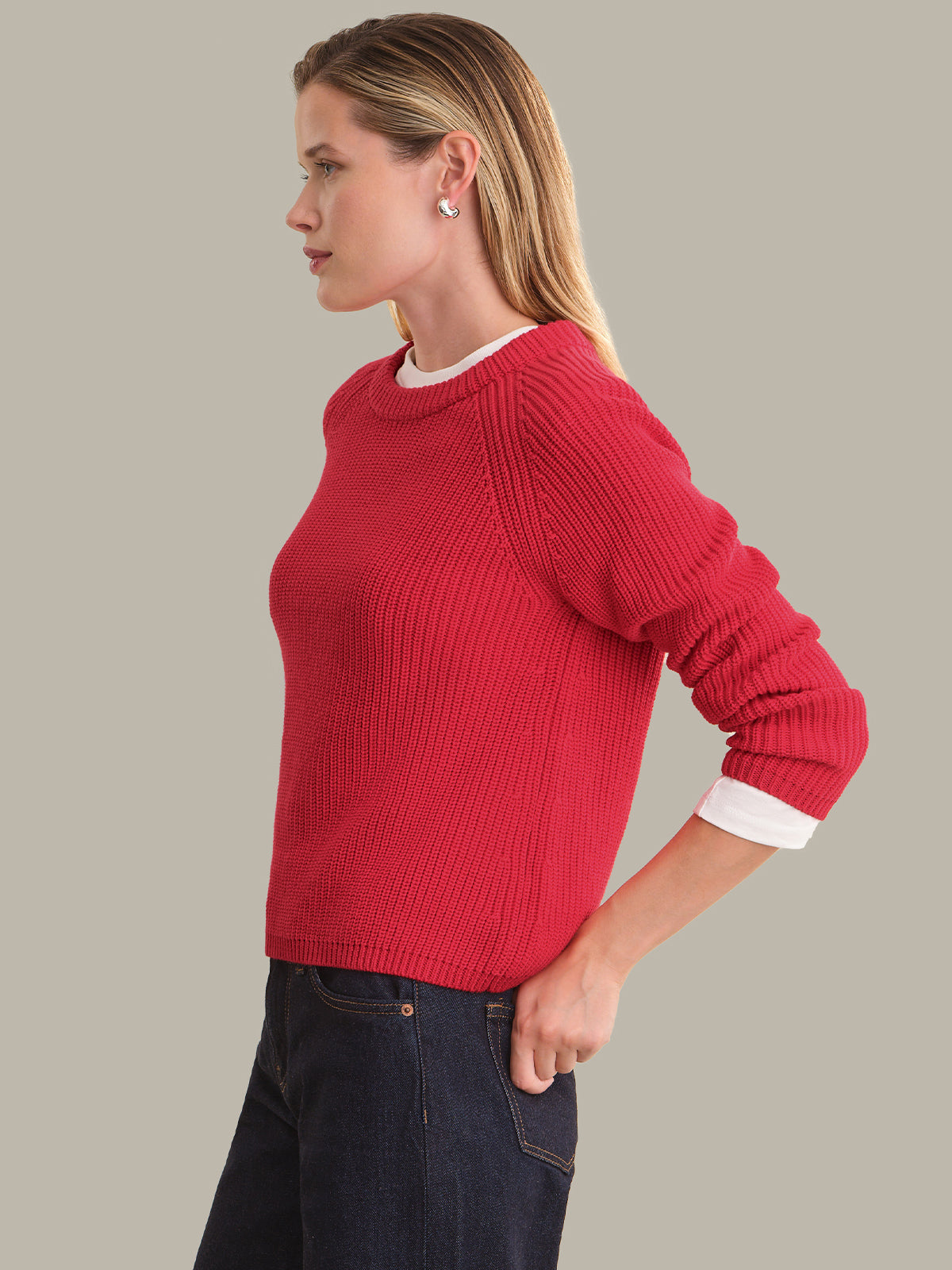 A woman in profile wearing a red ribbed knit sweater over a white undershirt and dark blue jeans, photographed against a solid light gray background.