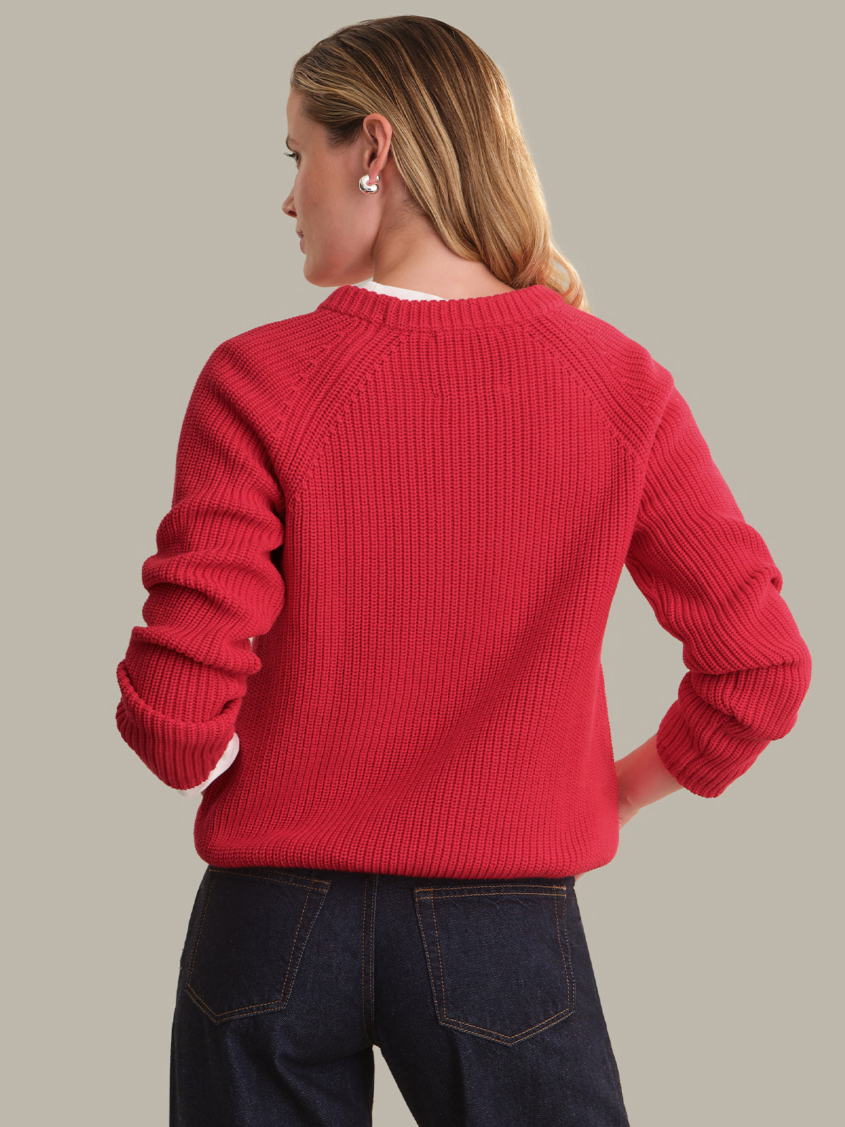 A woman shown from the back wearing a bright red ribbed knit sweater and dark blue jeans, photographed against a solid light gray studio background.