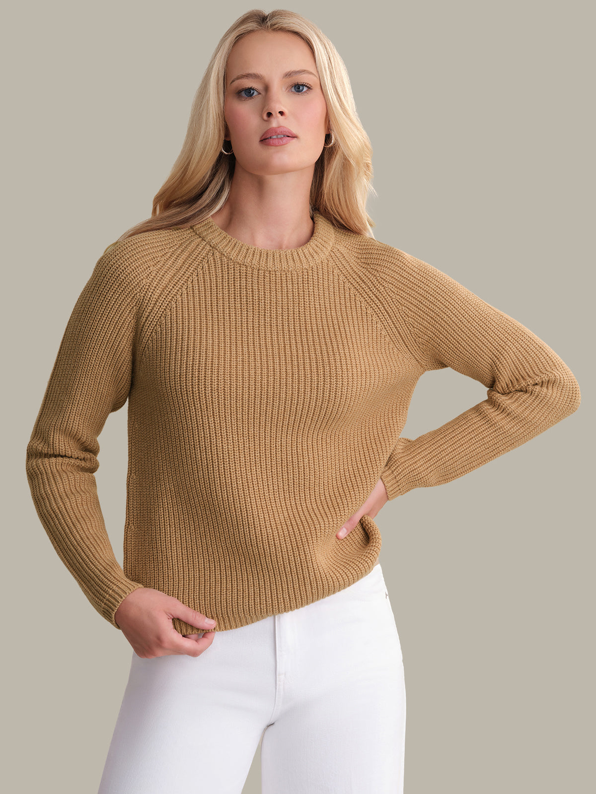 Woman wearing a beige sweater and white pants against a neutral background