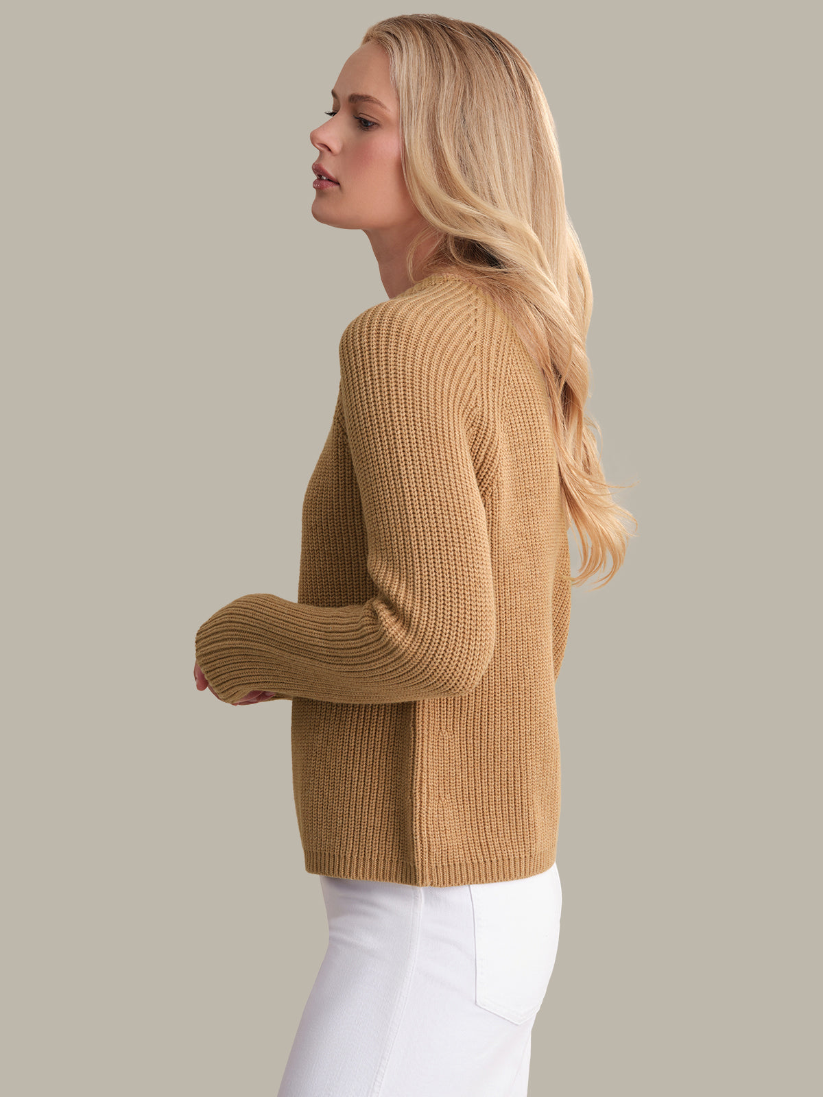 Woman wearing a beige sweater and white pants against a plain background