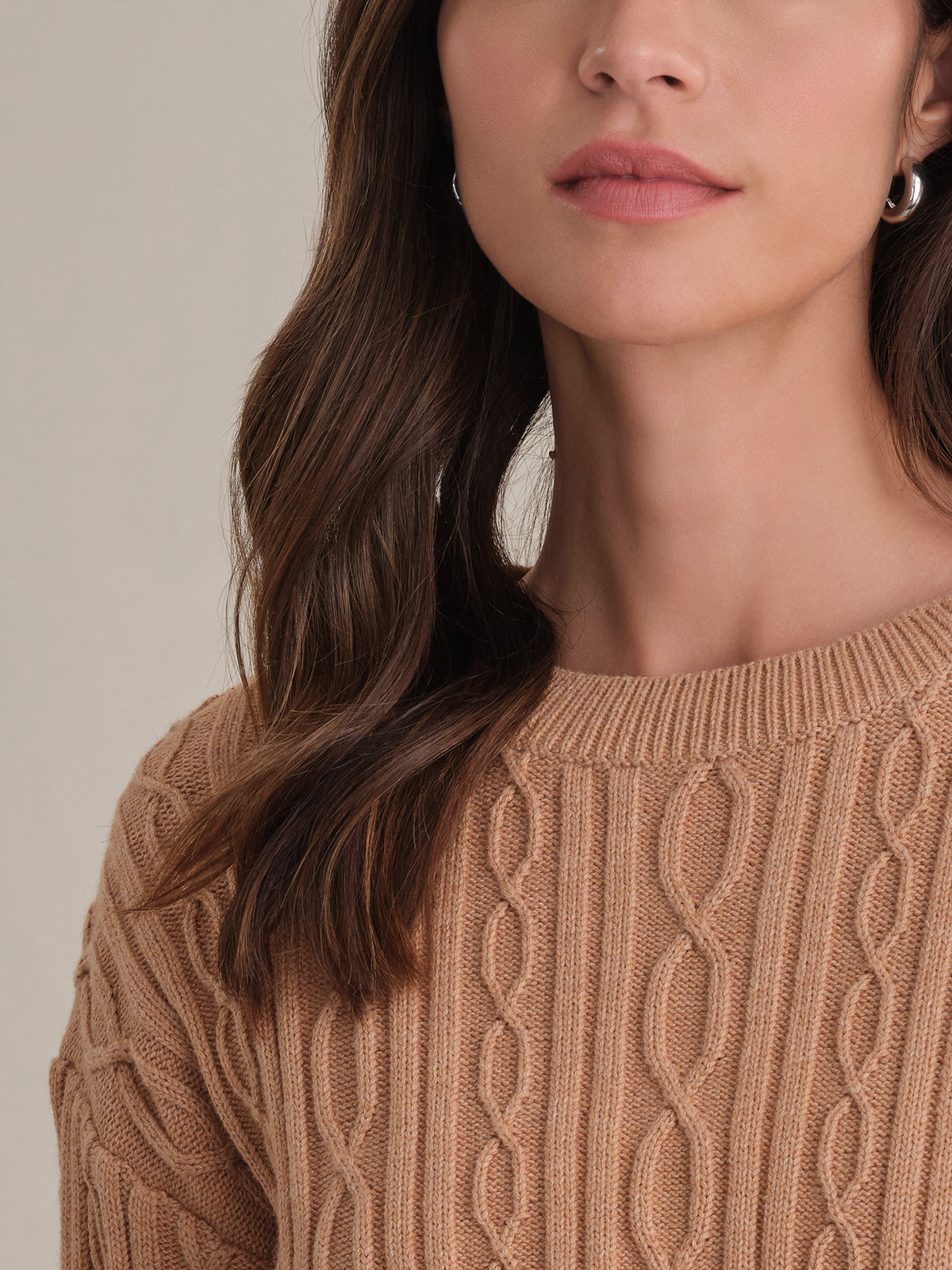 A close-up of a woman wearing a 525 America light brown Cable Cotton Sweater with a classic cable knit and crewneck. Only her lower face, neck, and upper chest are shown. Her brown hair is in loose waves, and she wears small hoop earrings.