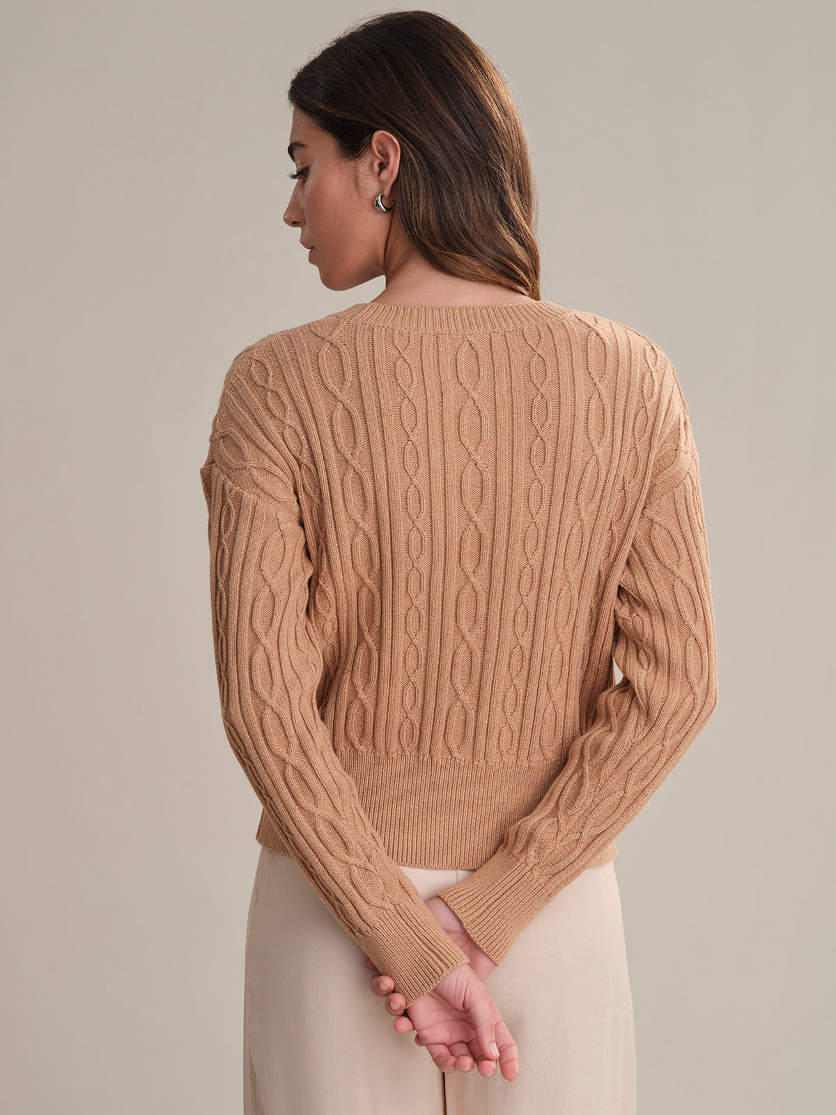A woman, her back to the camera, wears a tan 525 America Cable Cotton Sweater with a classic cable knit design and light-colored pants, hands clasped behind her against a plain background.