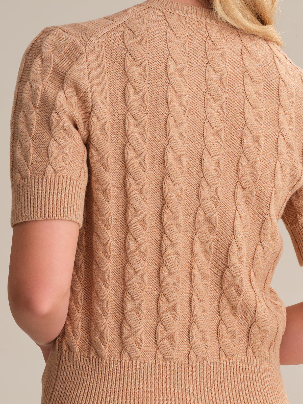 Someone is seen from the back wearing a short-sleeved tan cable knit sweater by 525 America. This cozy piece resembles a classic cable cardigan with ribbed hem and cuffs. The plain neutral background accentuates the sweaters seasonal shades and highlights the persons light blond hair.