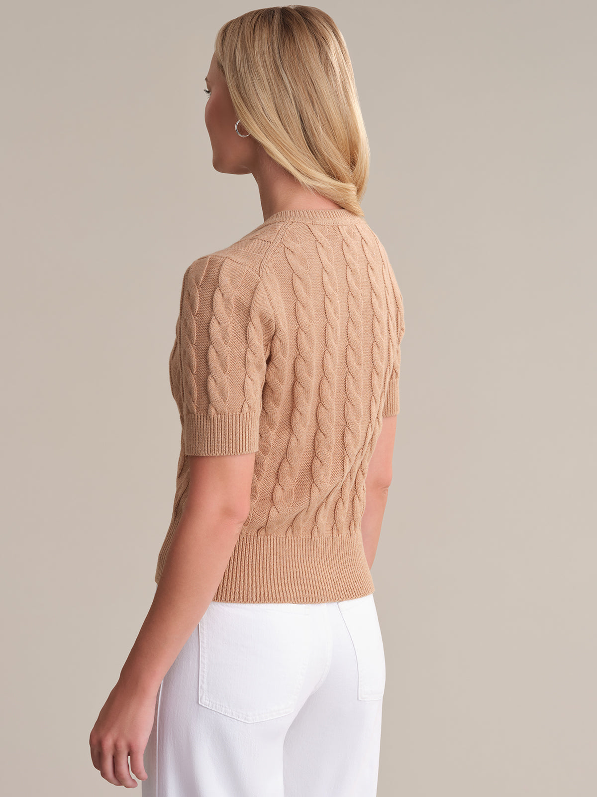 A person with long blonde hair wears a light brown, short-sleeved cable knit sweater from 525 America in seasonal colorways and white pants. They stand against a plain light gray background, turned slightly to the side while looking away from the camera.