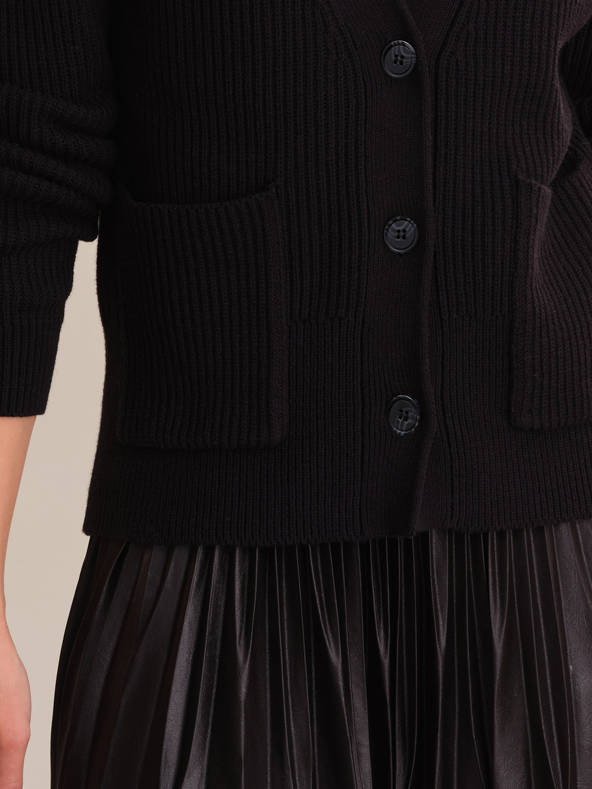 Close-up of a person in a 525 America black cotton cardigan with buttons and two pockets, paired with a pleated skirt. The v-neck design enhances the texture, and the bent arm highlights this elegant ensembles details.