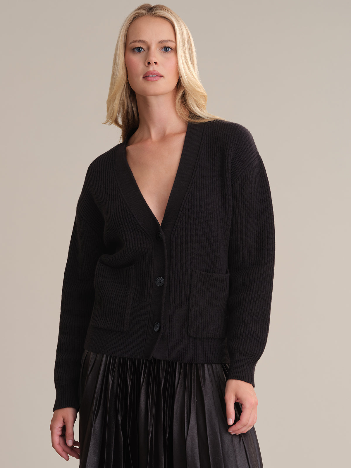 Someone with long blond hair is styled in a 525 America black deep V-neck cable knit cardigan, featuring buttons and front pockets, paired seamlessly with a pleated black skirt. The plain neutral background highlights the sophisticated ensemble.