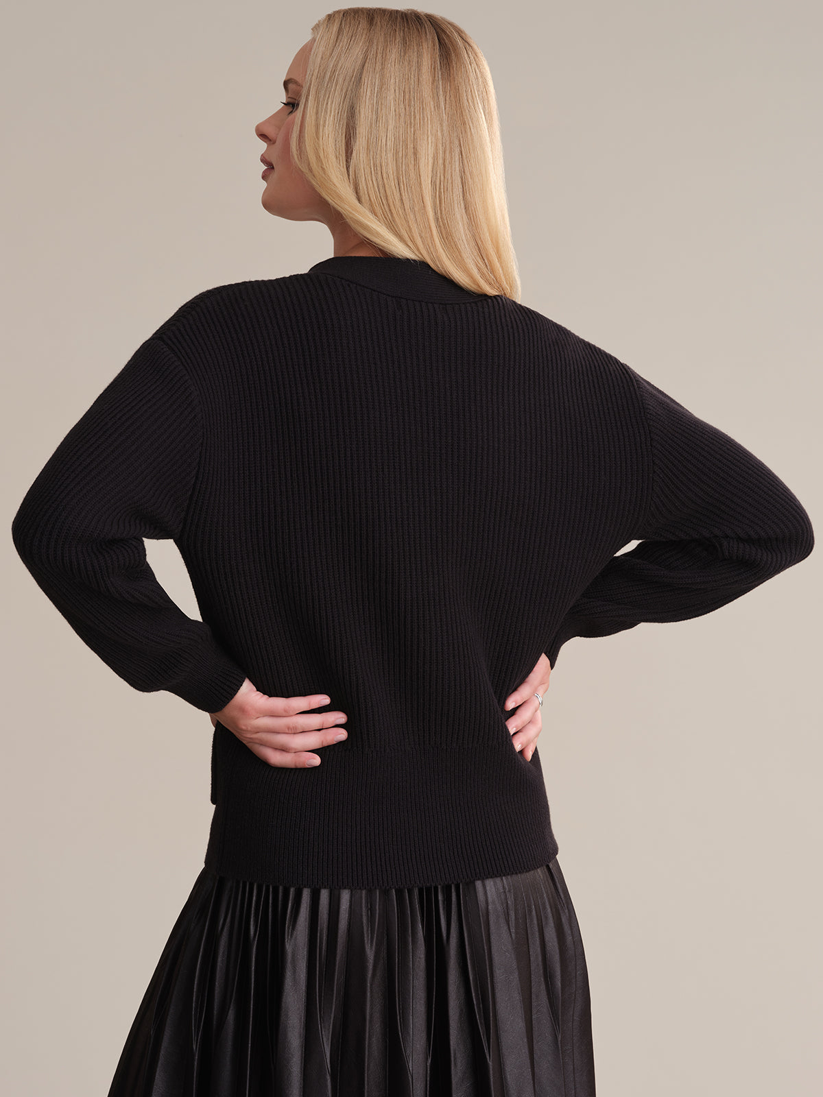 A person with long blonde hair stands back to the camera, wearing a stylish black ribbed sweater from 525 America, reminiscent of a classic cable knit cardigan, paired with a pleated black skirt. A plain beige background offers subtle contrast.