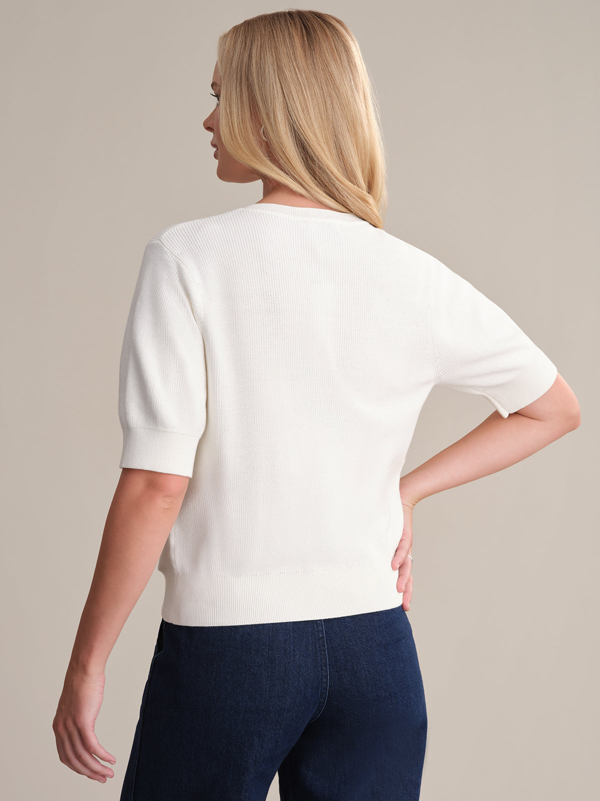 A person with light blonde hair is shown from the back, wearing a 525 America soft cotton Short Sleeve Puff Cardigan and dark blue jeans against a beige background. One arm is bent at the elbow and rests on their hip.