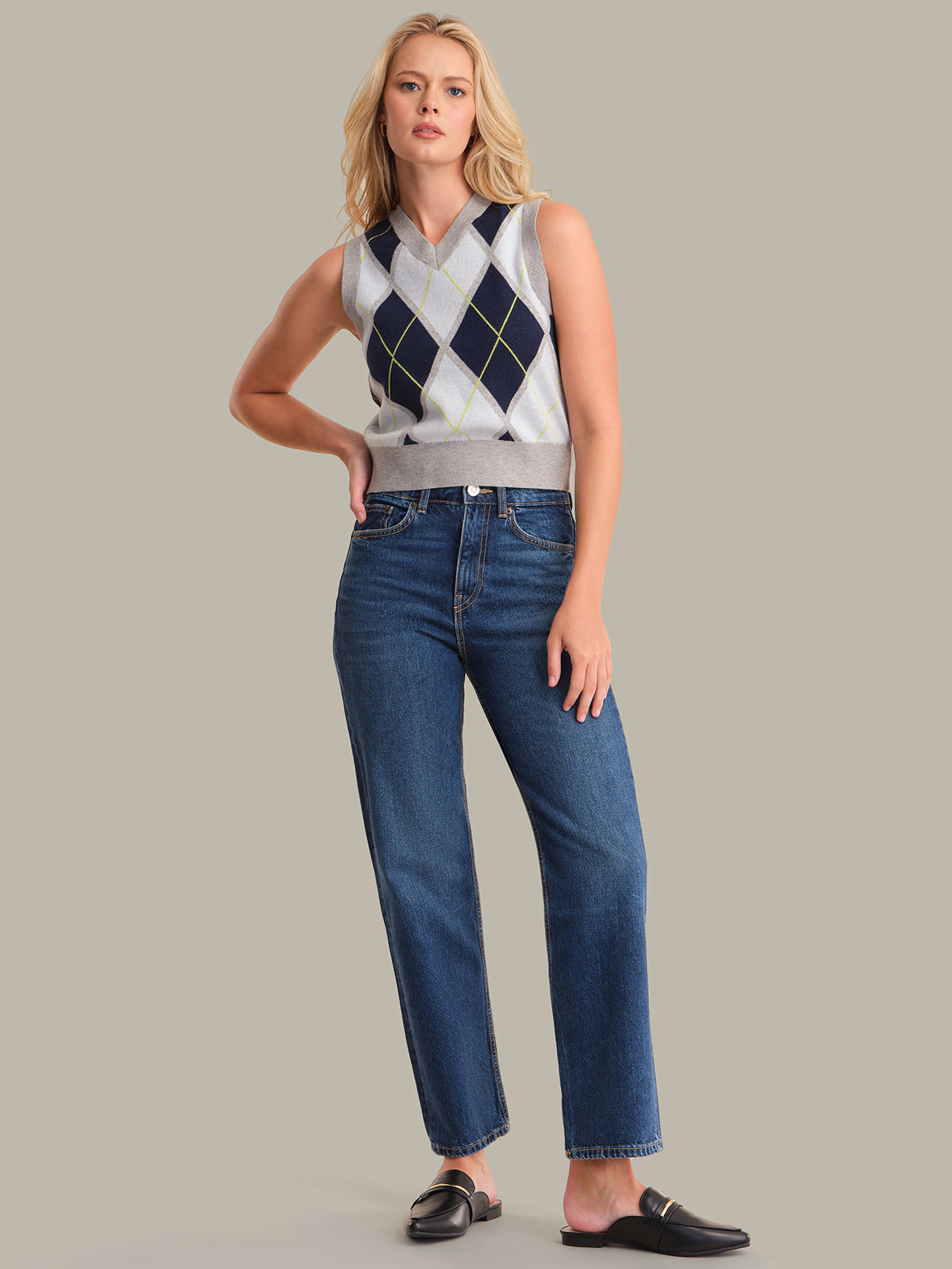 Woman wearing a sleeveless argyle sweater and blue jeans against a plain background