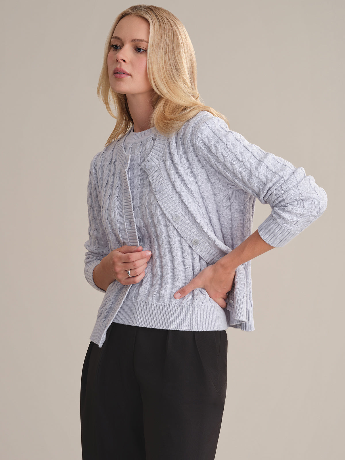 A woman with long blonde hair wears a 525 America cotton cable-knit cardigan over a matching top and black pants, standing sideways against a beige background with a neutral expression.