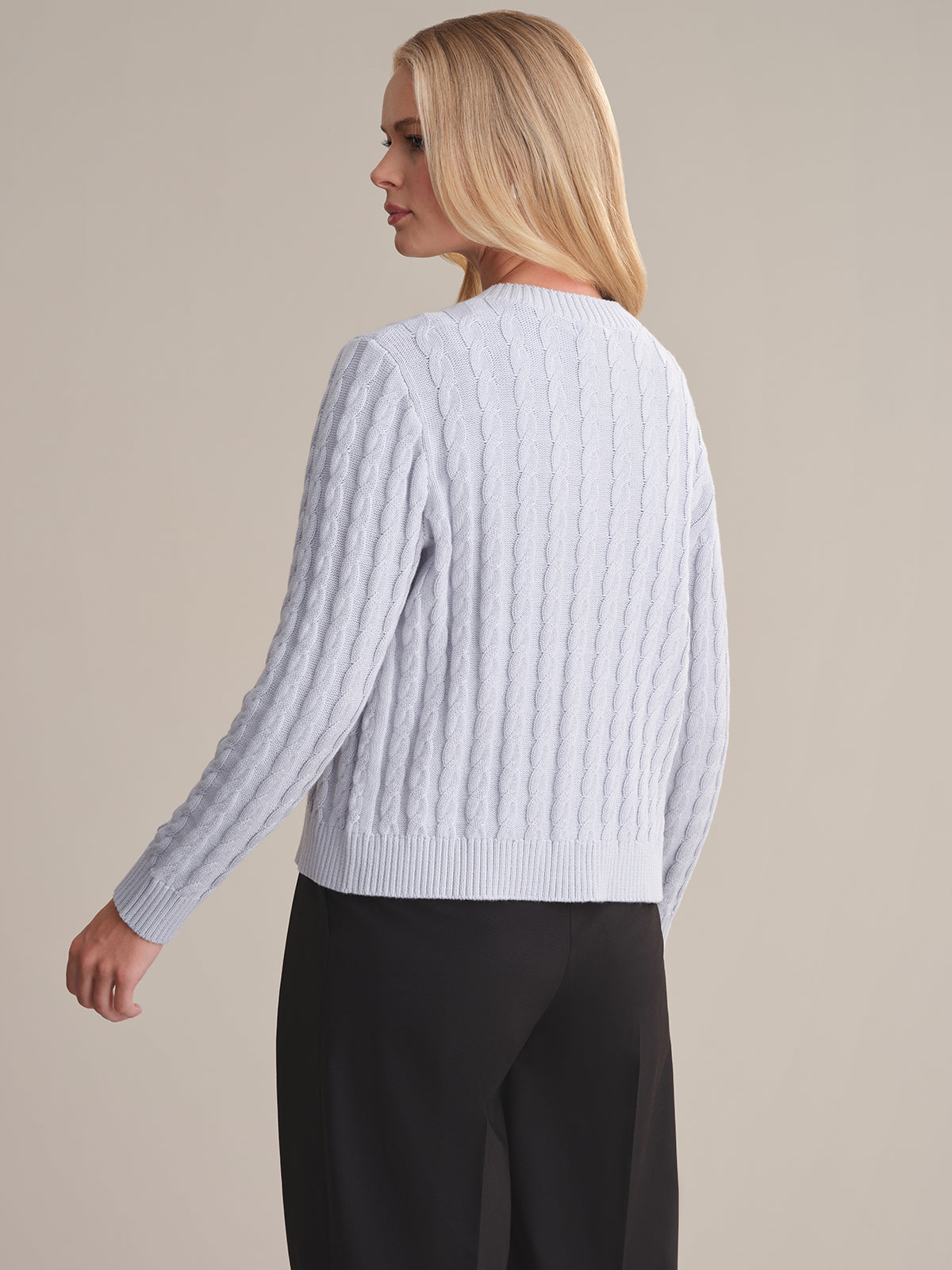 A person with long blonde hair stands side and back to the camera, wearing a 525 America light gray cable-knit sweater and black pants against a plain light background, showcasing the timeless style of the brand.